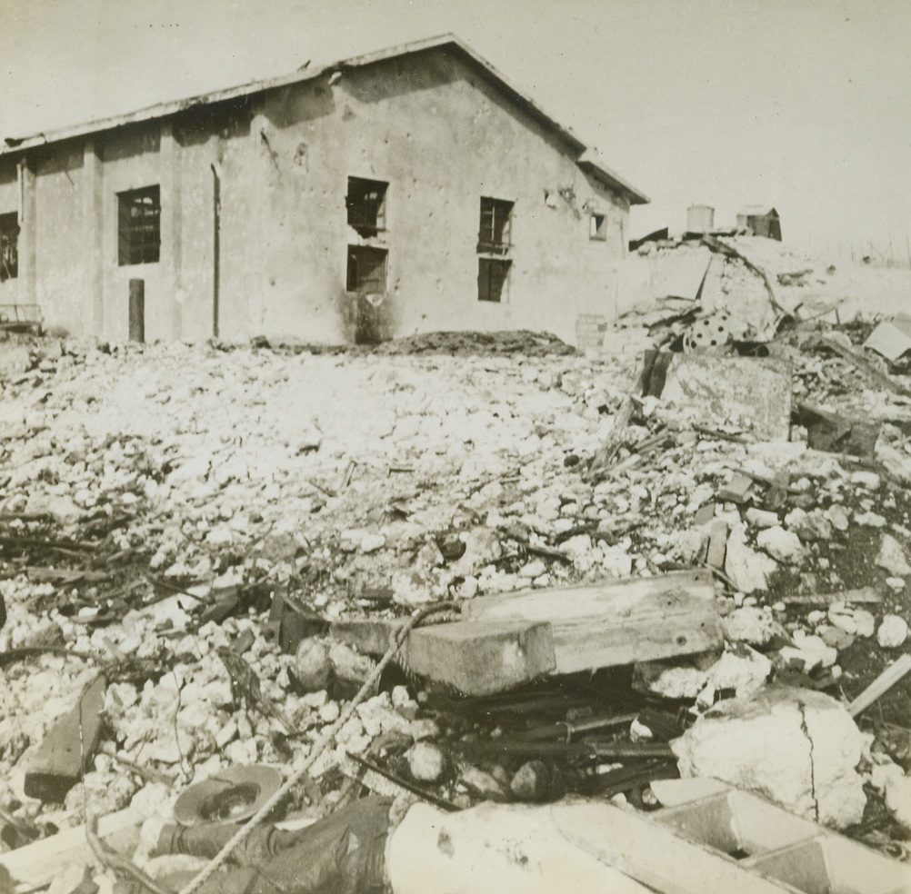 Wreckage – Man and Man Made, 3/14/1943. Tobruk, Libya – A dead Nazi soldier, washed ashore at Tobruk, lies half buried in the rubble of the city blasted and captured by the British 8th Army.  In the background, a bulled-scarred building has its foundation in a shambles that is evidence of the force of British fire power in the Autumn of 1942 when the Nazis were ousted from the Libyan port. Credit line (ACME);