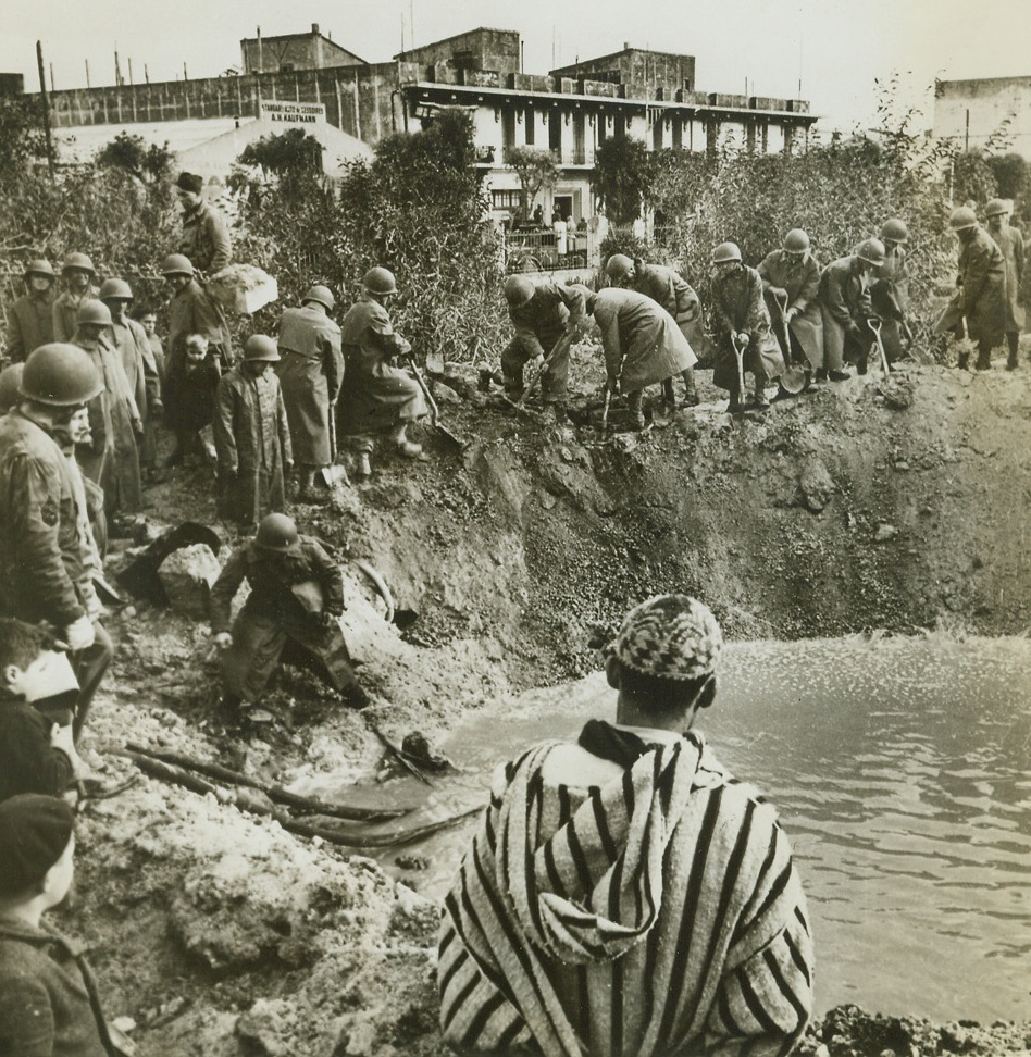 Quick Work, 3/7/1943. Casablanca – A native (foreground) watches as American soldiers fill in a bomb crater that was flooded by a broken water main which burst during a German bombing raid on Dec. 31.  Note that hoses pump out water even as the hole is being filled. Credit line (ACME);