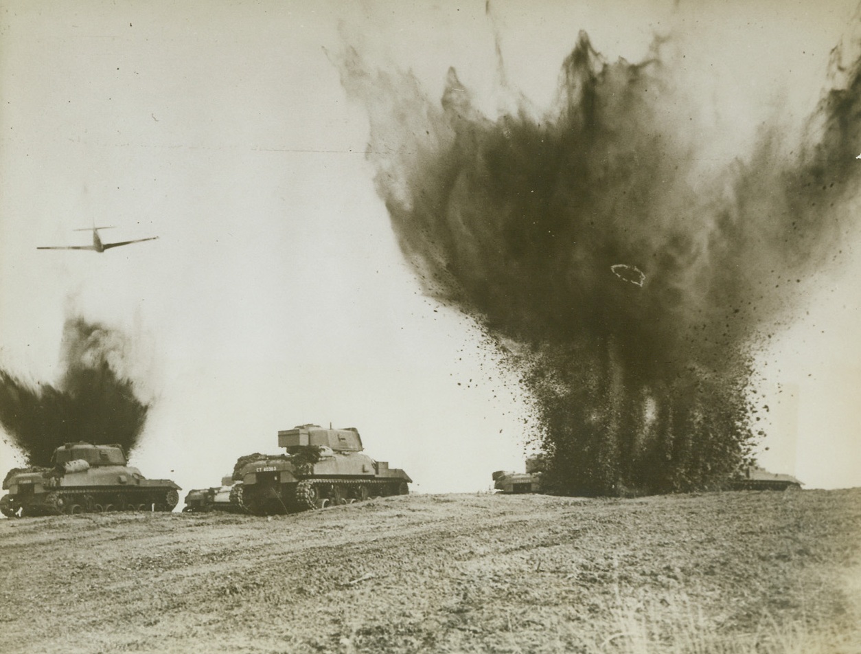 Dress Rehearsal, 1/14/1943. Somewhere in South England—This realistic dress rehearsal for their next big offensive against the enemy is carried out by Canadians stationed in England. Negotiating minefields, with mines exploding, Canadian tanks go through their grueling maneuvers with an aerial escort of mustang fighters constantly on the lookout for air attacks by the enemy. Passed by censor. Credit: ACME.;