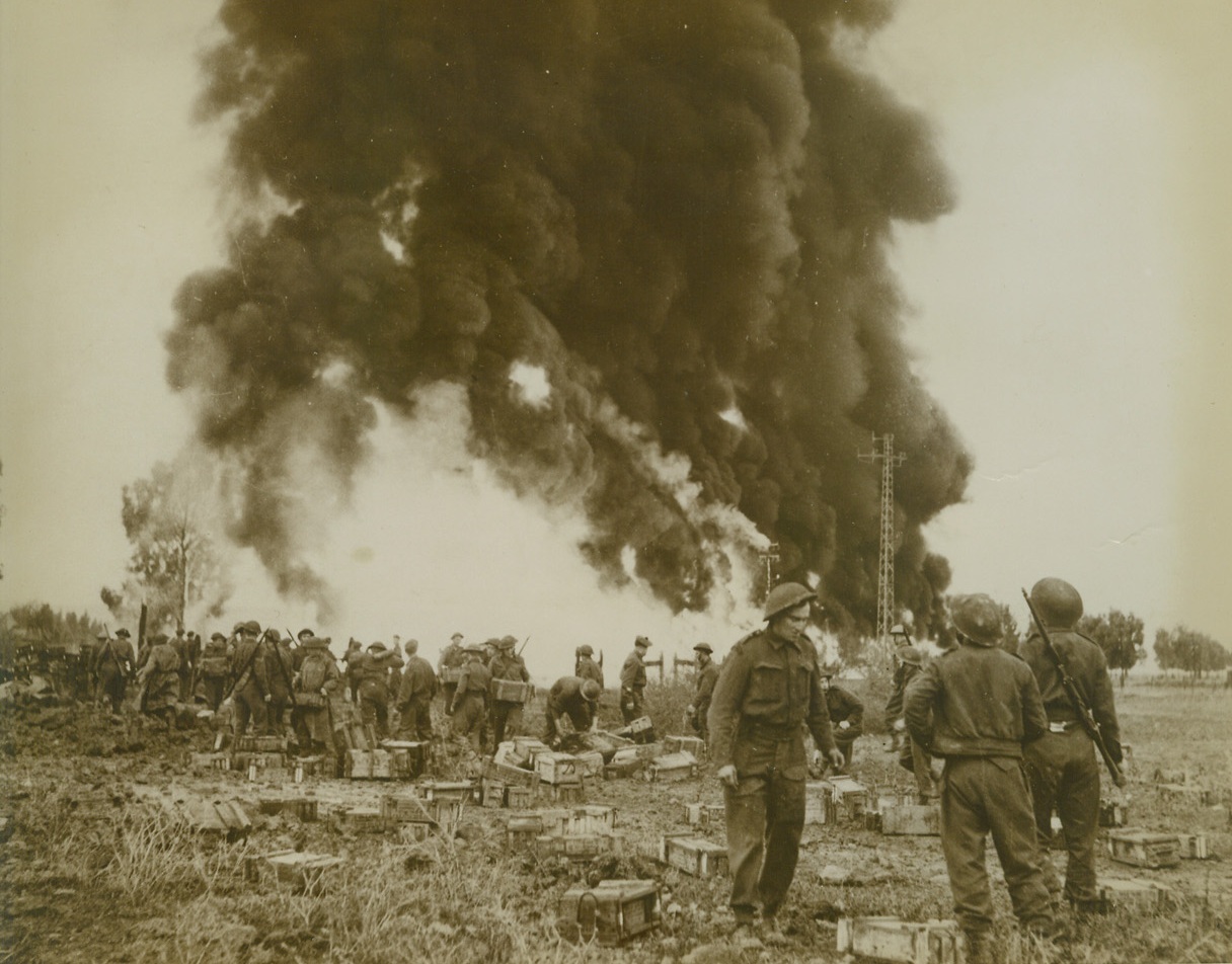 Flaming Backdrop, 9/15/1943. Tunis – With the smoke and flame of fires started by the Luftwaffe forming a backdrop, British soldiers speedily remove equipment from a bombed zone in Tunis.  Although this photo was made nearly a year ago, it has just been released by censors.Credit line – WP – (ACME);