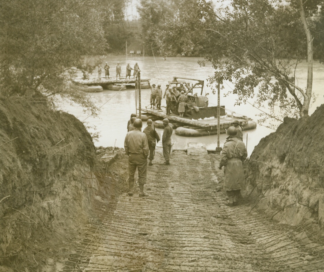 Nazi Hopes Drowned In The Volturno, 10/25/1943. ITALY—When 5th Army troops splashed, paddled or swam across the Volturno, more than a river was lost to the Germans. The Nazis learned in blood that their natural barrier defense system wouldn’t work. To further consolidate this message of Axis defeat, crack American engineers immediately floated a pontoon bridge across the shallow river so that heavy Allied equipment could rumble on the heels of the advanced infantry forces who had begun the job of dislodging the enemy. These photos tell the story of that bridge which, in reality, may be called a road to victory in Italy for the Allies.  NEW YORK BUREAU A bulldozer (foreground) is ferried across the Volturno while (background) infantrymen of the 5th Army make the crossing via pontoon raft. The building of the bridge eliminated the necessity of the slow ferries for transporting heavy equipment. Credit: Acme photo by Bert Brandt, War Pool Corresponden;