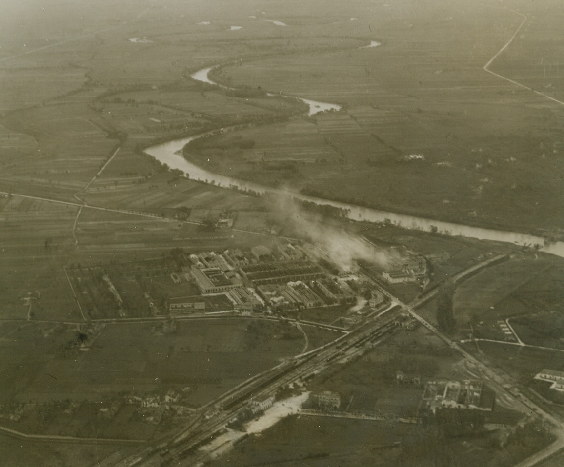 ARTILLERY DUEL ACROSS THE VOLTURNO, 10/25/1943. ITALY—White smoke marks an Axis hit on the British-held town of Capua, during the fierce battle for the Volturno. In the background, that river follows a corkscrew path which was once spanned by bridges destroyed by the Germans. This photo was made from an artillery reconnaissance Piper Cub flying between 2,000 and 3,000 feet over Axis and Allied lines. Credit: Acme photo by Charles Seawood, War Pool Correspondent;