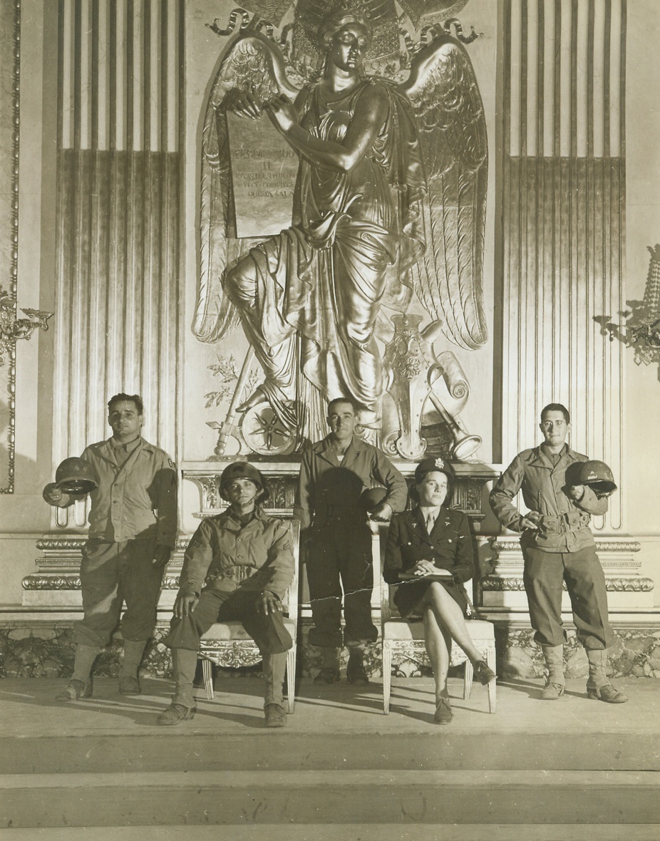 “Royal” Tintype, 11/17/1943. NAPLES—Cpl. Joseph Barber, Girard, Ohio, is “King” and Army nurse Lt. Maryellen McCutcheon, Birmingham, Mich., acts as “Queen” while Pfc. William Dato, Hoboken, N.J.; Pfc. William Montegomery, Detroit, Mich.; and Cpl. Thomas Shaw, New York; are the “courtiers” in this little tableau snapped in the throne room of one of the royal palaces in Naples. The gold throne was removed to another part of the palace for safekeeping, so chairs have to serve the “royal” pair. Credit: ACME photo by Bert Brandt, War Picture Pool Correspondent.;