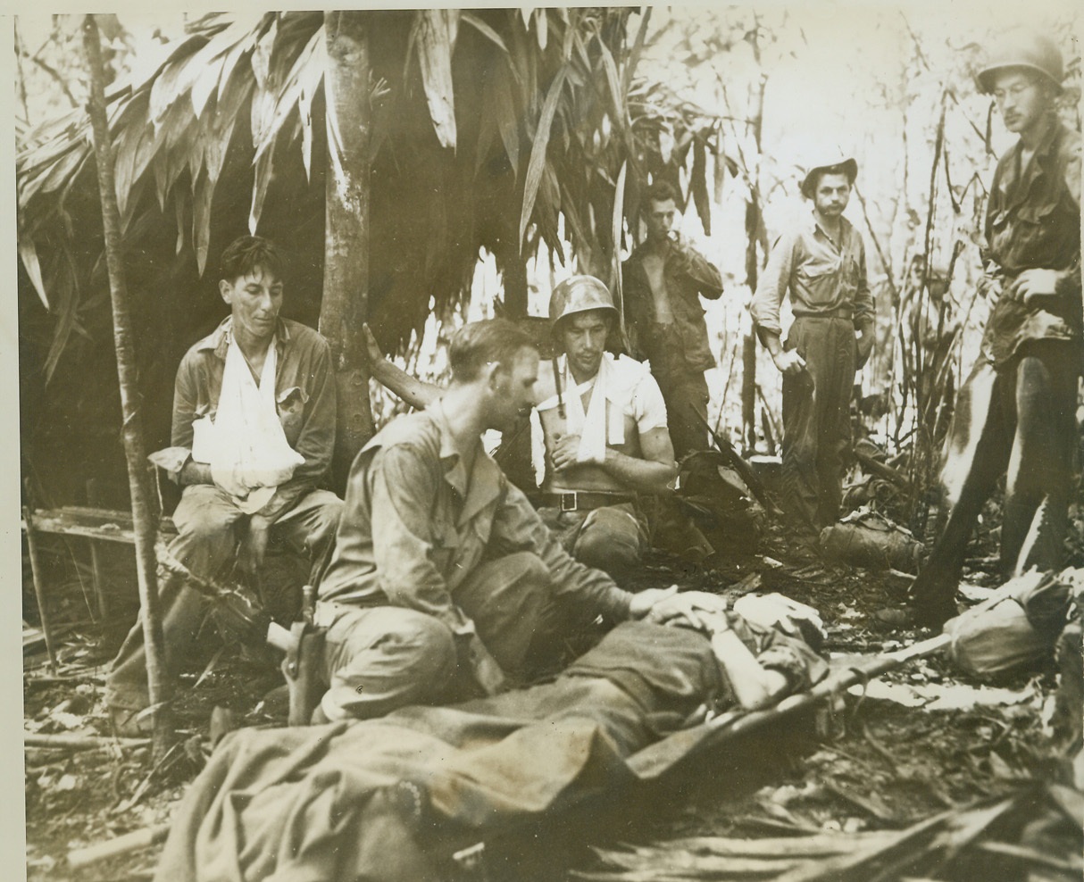 Wounded on New Georgia, 11/24/1943. NEW GEORGIA ISLAND -- Exhausted, the lines in their faces revealing the strain under which they've been fighting, these Yanks stop to rest in a deserted Jap bivouac, from which the boys chased the enemy. Those who escaped injury watch a Medical Corps officer treat an unidentified wounded man. Watching, too, are Sgt. Morris Hain (extreme left), whose arm is in a sling, and Cpl. Hollis Templeton, whose left shoulder is bandaged. Credit: (Signal Corps Photo from ACME);