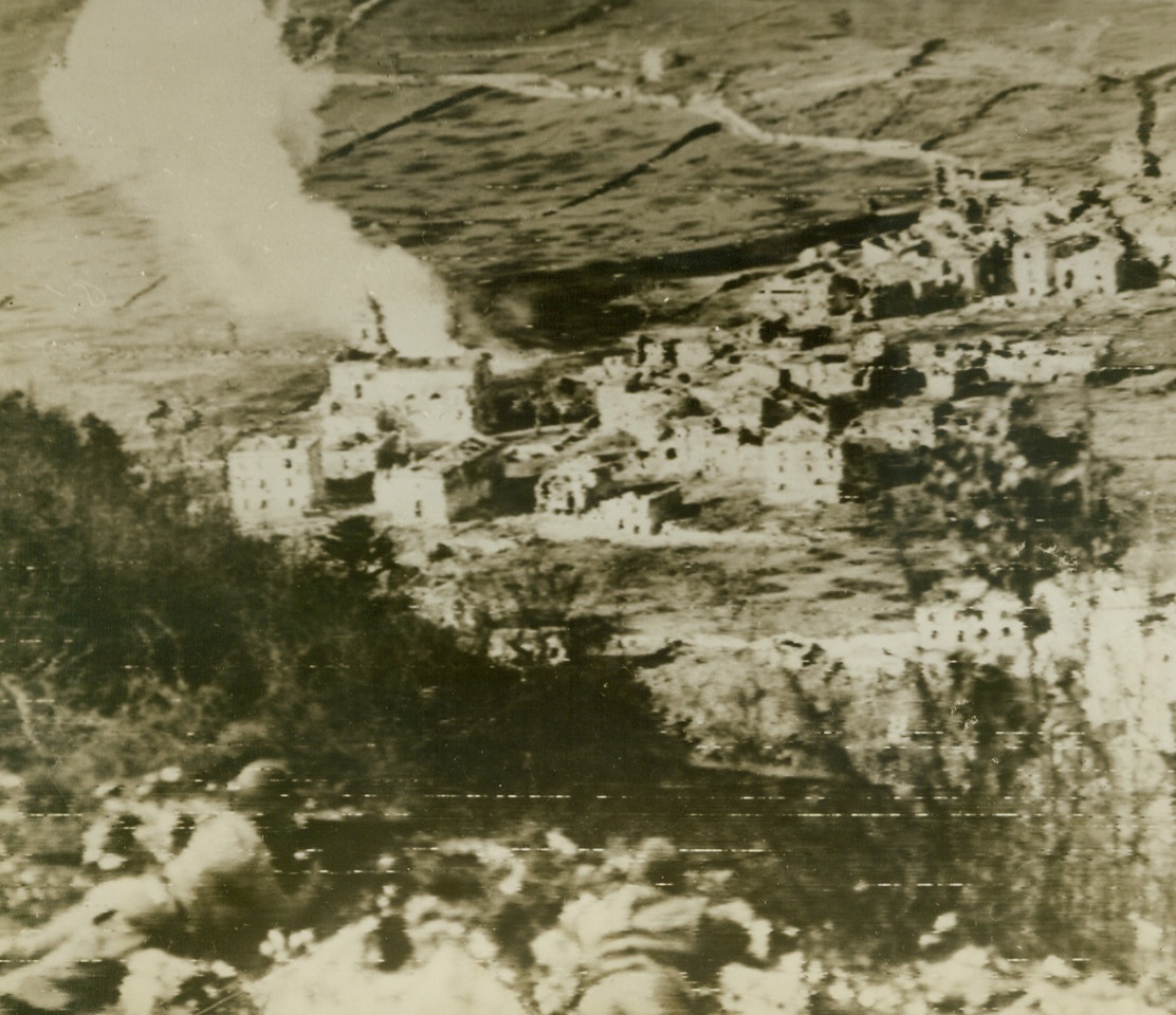 Dropping Shells on Nazi Headquartersf, 12/16/1943. CONCA CASALE, ITALY – A towering column of white smoke marks an exploding American shell, just beyond the church (note steeple), which houses the German headquarters in Conca Casale, just North of Venafro. In foreground, (photo above), U.S. snipers lie ready to pick off any of the Nazis who try to escape from the bombarded building.  Credit Line (ACME Photo by Bert Brandt for the War Picture Pool transmitted by Signal Corps Radiotelephoto);