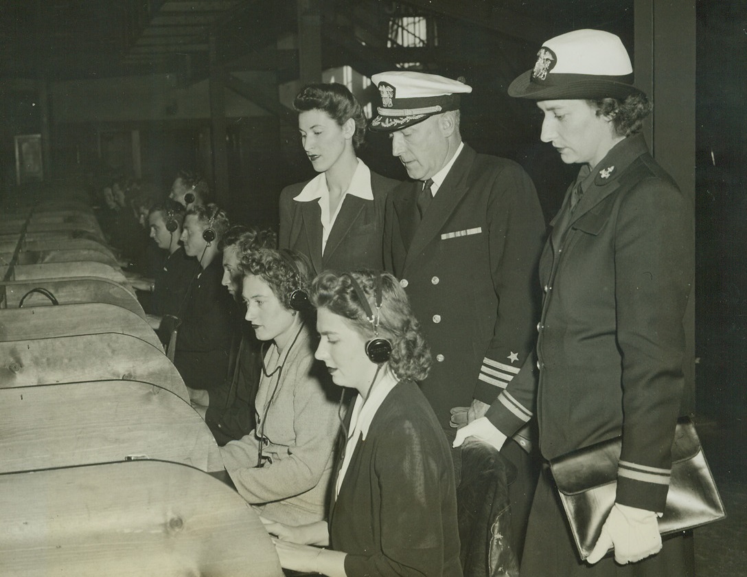 Noncommissioned WAVES Go Into Training, 10/11/1942. Madison, Wisc. – Comm. Charles P. Greene, U.S.N., former Assistant Chief of Staff of the 5th Naval District, Norfolk, Va., and new Commanding Officer of Naval Training Schools at the University of Wisconsin, Madison, Wisc., watching the first of 480 noncommissioned WAVES try their hands at typewriters and earphones in the radio classroom, where they will get a three-month course in radio communication. Left to right, standing: Miss Betty Kam, Clasonpoint, N.Y., a recruit; Commander Greene; Lt. Jenny Turnbull, Washington, D.C. Seated: Miss Jeanne Grey, Oakland, Calf.; Miss Lollie Catts, St. Louis, Mo. Credit: ACME;