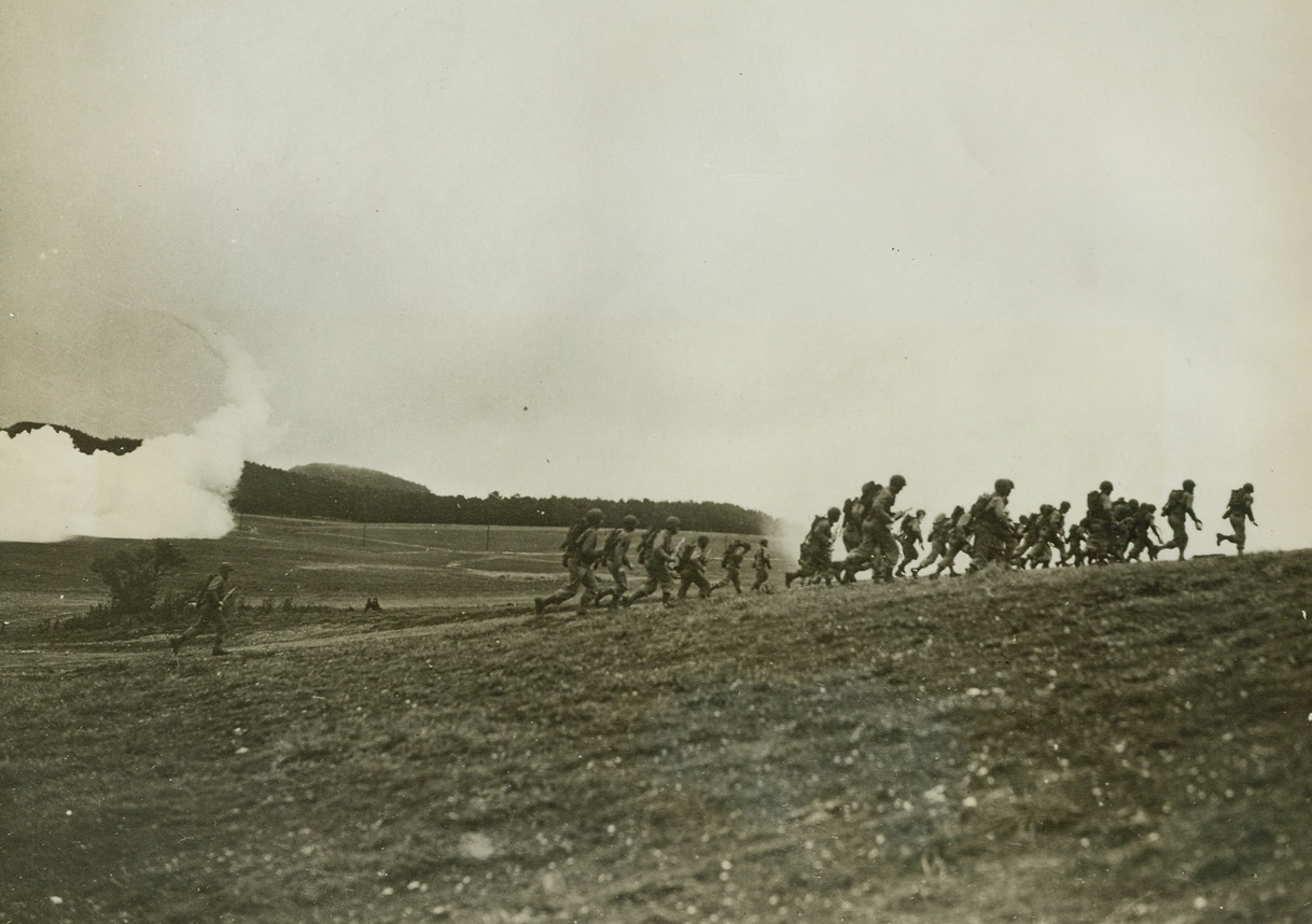 Smoke Screen Hides Advance, 10/13/1942. England – Troops advancing to the crest of a hill make splendid targets for the defenders when they are silhouetted against the sky. To prevent this, low-flying planes lay smoke screens enabling the attackers to meet the foe on a more even plane. U.S. forces in Britain, holding their first maneuvers that stressed the use of chemical warfare, are shown making an advance under such condition. During the maneuvers, an infantry unit was “attacked” by “enemy” planes that dropped smoke bombs to break up their fire-power, and also simulated mustard gas. Mechanized jeeps and scout cars were thrown into the attack, and flame-throwers were used to clean out strong points to give the soldiers a taste of modern fighting. Passed by censor. Credit: ACME;