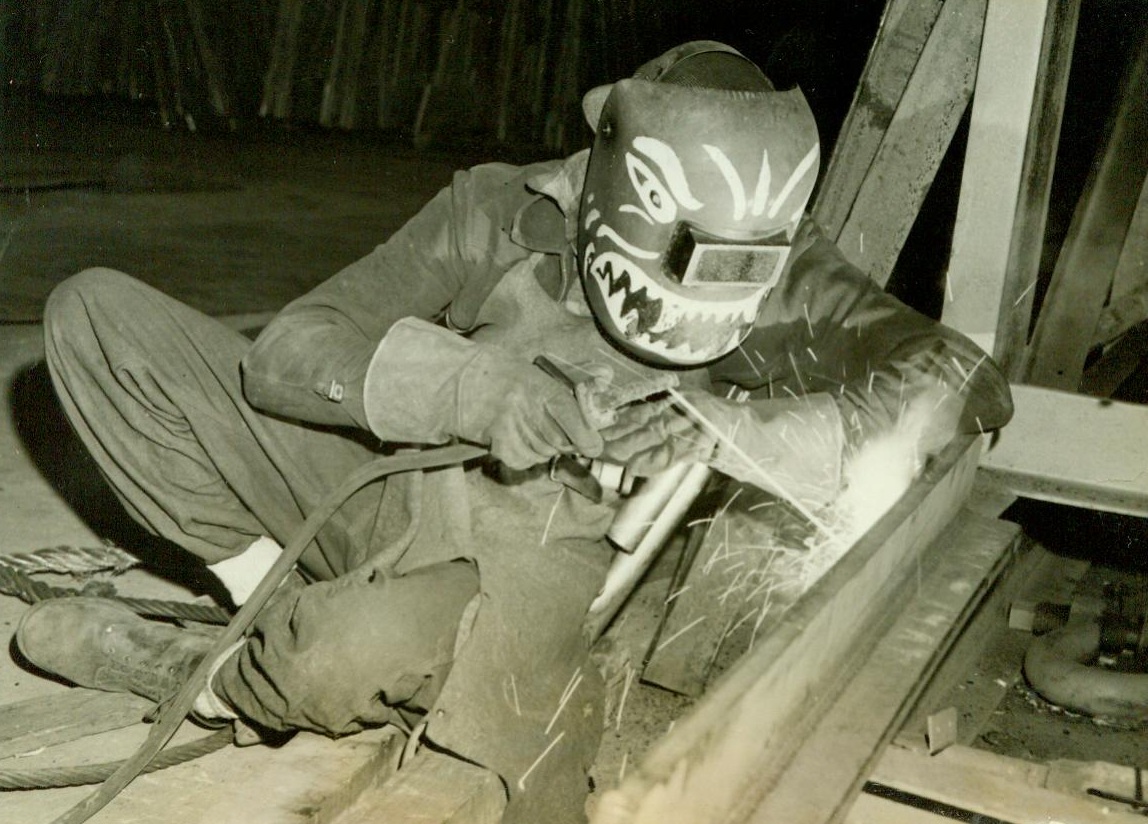 Pre-fabricated Ships Built 4 Miles From Ways, 12/12/1942. CHICAGO – Traditionally, laying of the keel marks the beginning of a new ship, but under a system developed by the Pullman-Standard Car Manufacturing Company, naval patrol bessels are being constructed in 14 completed sections simultaneously, the keel being a part of each section. Each of the 14 sections is assembled complete with deck plates, bulkheads, and much of the interior fittings, in a building of the Pullman Plant, Chicago, and carried on flat cars to the ways, four miles distant. At the ways, the center and stern sections are assembled first, because they require more interior fitting. This fitting is done while the bow sections are being put one time, have been built on reclaiming land at the end of Lake Calumet, on the far south side of Chicago. PHOTO SHOWS one of the welders his mask painted to resemble the Flying Tiger insignia, working on a pre-fabricated section. CREDIT (ACME) 12-12-42;