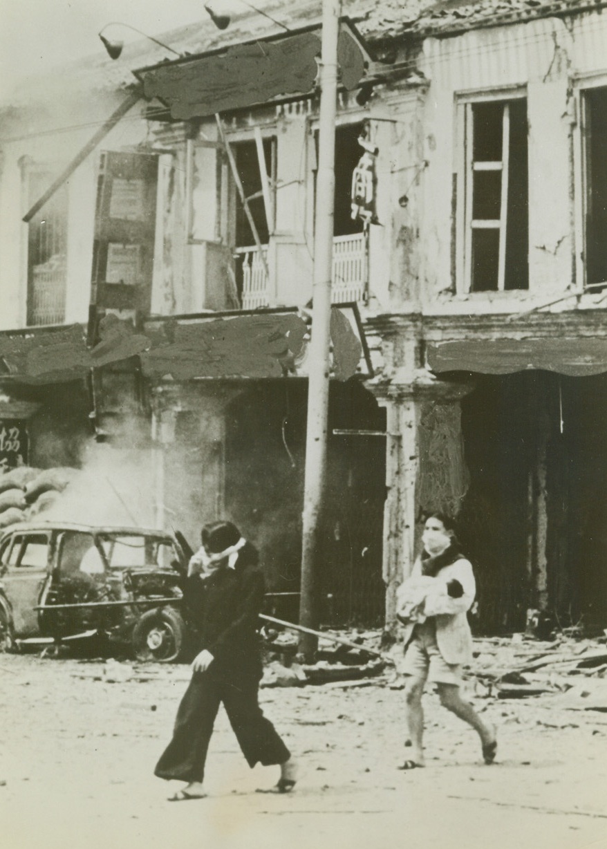 Last Days of Singapore, 3/17/1942. Singapore – Handkerchiefs tied across their faces to serve as improvised masks against the sting of explosion gases, natives of Singapore, one of whom carries a baby in her arms, are rushing from a bombed and burning building.  The last days of the Great British bastion were a nightmare of incessant bombings, fires and the tortures that came through a lack of water after the Japanese cut off the water supply to the city. Credit line (ACME);