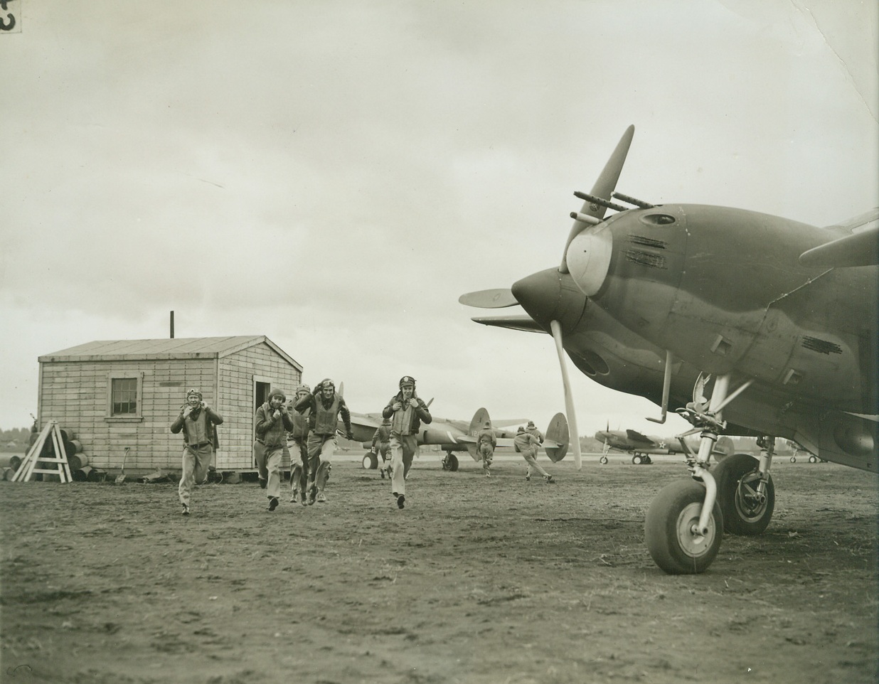 No Title. 5/8/1942. “Somewhere in the Pacific Northwest” one of Uncle Sam’s interceptor pursuit squadrons is shown going through a typical day of war time activities as they train to knock the enemy out of the sky should he make an attack on our West coast.  From dawn to dusk the entire squadron is on the alert as a matter of general routing, but during the rest of the 24 hours each day, smaller units take turns standing by while the rest of the men in the squadron study, service their fleet pursuit ships, play or catch up on their sleep. New York Bureau Pilots can be seen dashing to their planes from an alert shed just after receiving the “scramble order”.  The sheds are conveniently located on the field so the pilots can reach their planes in 15 seconds.  Ground crews warm the motors of the ships every hour, like clockwork, so the planes are ready for an immediate take-off. Credit line (ACME);