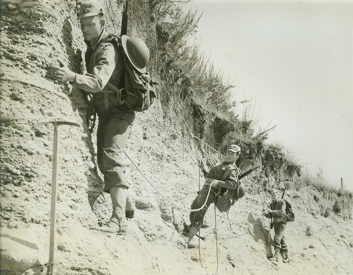 Ski Troops in Winter-Mountaineers in Summer, 8/16/1942. PACIFIC NORTHWEST – A new branch has been added to Uncle Sam’s Army – a full-fledged “Mountain Regiment”, with 1,400 men who are undergoing a rigid and specialized training program. Last winter these same men formed the first organized ski troop battalion in the U.S., but now that the ice and snow has given way to summer heat and dust they are being developed into a regiment intended as a nucleus for similar units of mountain infantry. The regiment’s commanding officer, Col. O.S. Rolph, inaugurated the ski patrol and today personally supervises every phase of the thorough training of the “Mountaineer Infantry”. These photos, taken at the Mountaineering Training Area, show some phases of the training. New York Bureau Mountaineers edge carefully along the face of a rock wall as they complete their basic training and prepare for the second phase and more rigorous training in a wild and mountainous area.;