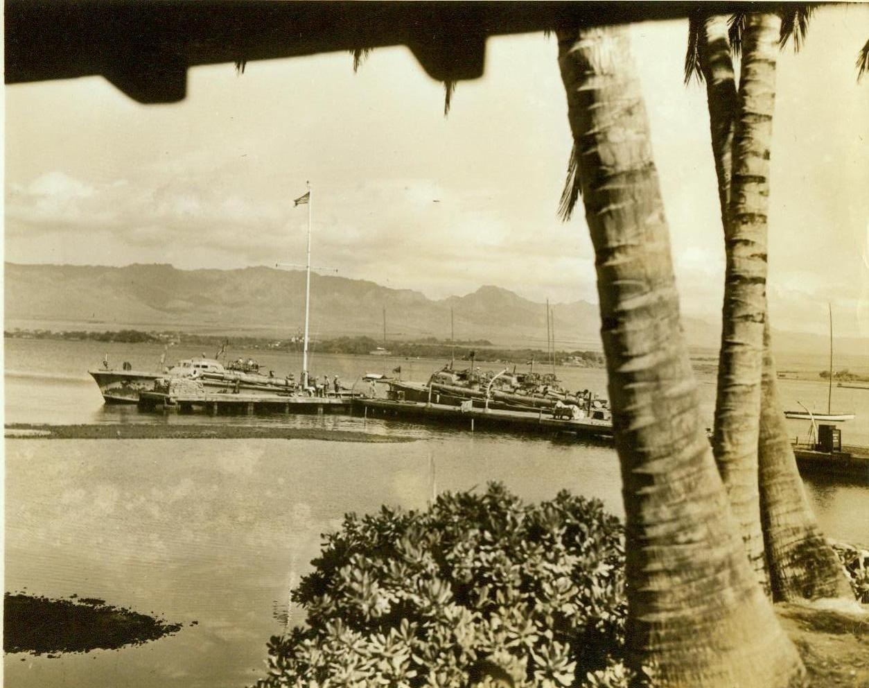 HAWAII’S GOT STINGERS NOW, 8/1/1943. HONOLULU – A palm-fringed, tropical lagoon is headquarters for a nest of sea-going hornets whose stings are deadly to any lurking Jap submarine or surface vessel off Oahu’s shores. It’s the home of the sturdy, speedy little PT fleet – the Navy’s torpedo boats that have recently joined land, sea and air offensives in Hawaii. In a former yacht clubhouse set beneath cocoa palms and huge Kamani trees, the officers and men of the fleet relax while their craft undergo check-ups and repairs, awaiting the time when they will put out to sea—for a day’s or month’s cruise—to “put the fish into an Axis ship”. Here are typical scenes at the secret base. New York Bureau These “sub-dusters” are tied up to their docks, awaiting repairs in the calm waters of the lagoon at the secret torpedo boat base. (Passed censors). Credit: ACME;