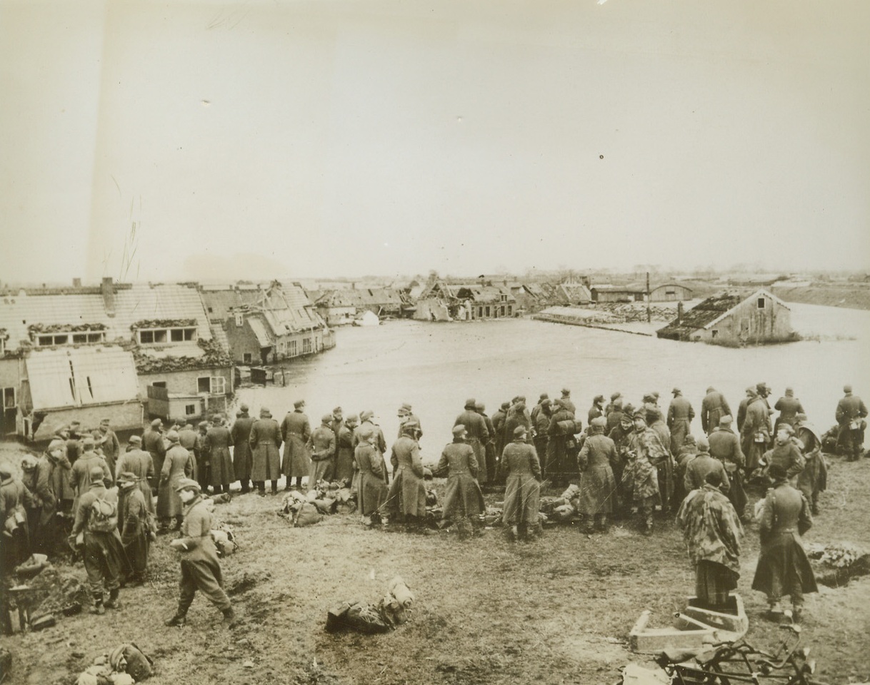 NAZI PRISONERS IN FLOODED DUTCH TOWN, 11/14/1944. HOLLAND—Some of the thousands of Nazis taken prisoner at Middleburg stand on high ground waiting for Allied vessels to carry them across the canal to prison camps. Town in background is almost completely submerged as the sea poured through breaks in dykes opened by Nazi demolition experts. Credit (British Official Photo from ACME);