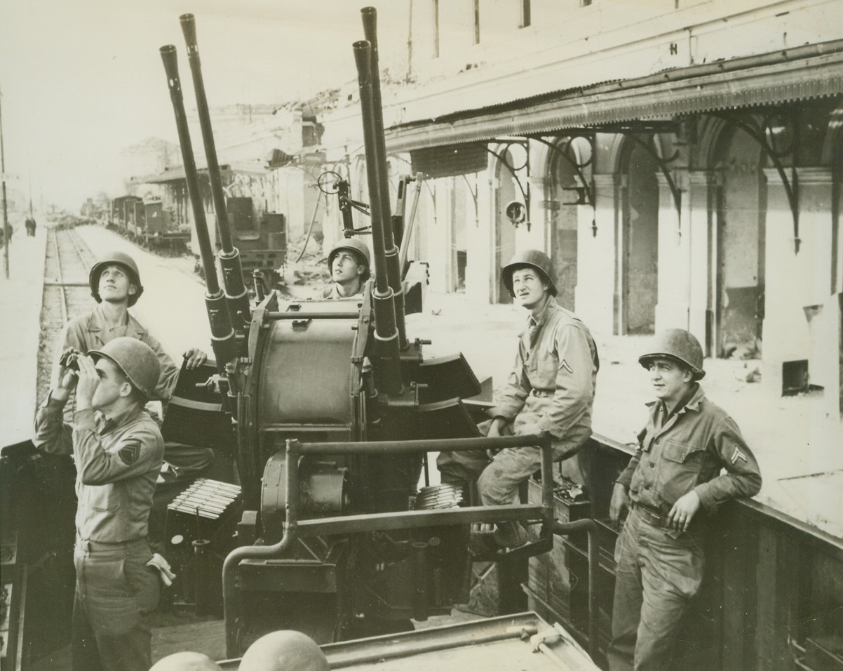 Triple Play—Hitler, To Musso, To Yanks, 1/25/1944. Naples, Italy—This deluxe, super armored and armed railway car was presented to Mussolini by Hitler (P.S.: In the “Good Old Days”). It was built in Germany, has luxurious living quarters in the center of the car and quadruple gun mounts at each end. As the Germans were chased out of the Naples area, they tried to destroy it but the Yanks were too quick for them. Now it is a mobile anti-aircraft gun mount and is manned by an American crew of 10, who live in the fancy quarters meant for Mussolini. Here (Left to Right, clockwise around gun), are: Sgt. John T. Adams, of ST. Louis, MO; Pvt. Joseph N. Schan, Pardeesville PA.; S. Lolrich, Denver, CO; and Cpl. Albert E. Nickel of Chicago, ILL. The men show here during a practice alert, all members of an airborne machine gun battalion. Credit: OWI Radiophoto via U.S. Signal Corps from ACME;