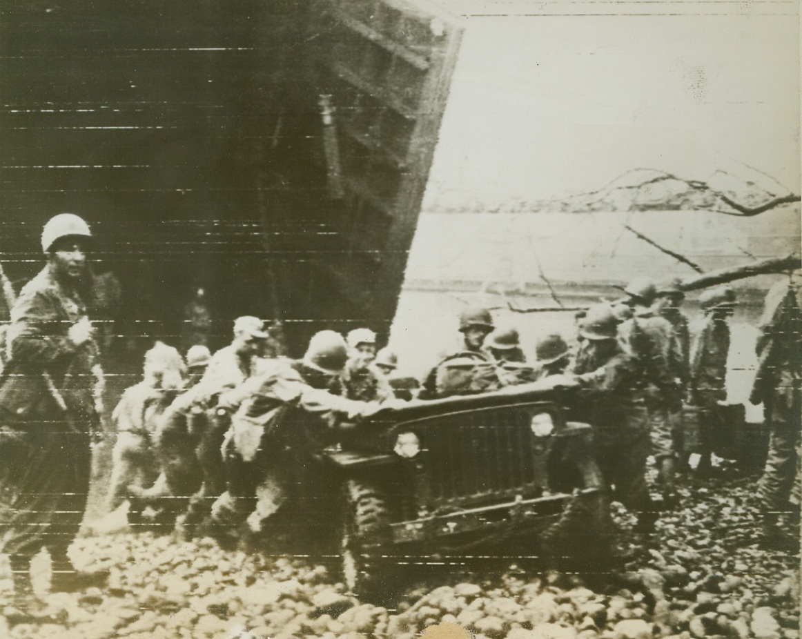 Heave – Ho, 1/6/1944. Saidor, New Guinea --  Fresh off an LST, this jeep makes a bumpy trip up the beach at Saidor, New Guinea.  Just landed, a group of American soldiers escorts the tiny car over the rough path to newly-laid road mats.Credit (Signal Corps radio telephoto from ACME);