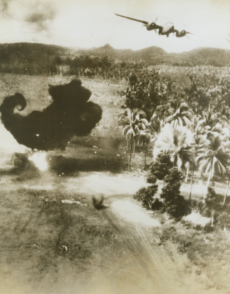 Treetop “Call” for Japs, 1/25/1944. A B-25 Mitchell bomber of the U.S. Army 5th Air Force, comes in at treetop level to blast installations, while a Jap plane burns fiercely (left) on the fighter strip at Alexishafen, above Madang on the North coast of New Guinea, during a recent raid.  Credit line (U.S. Army Air Force photo from ACME);