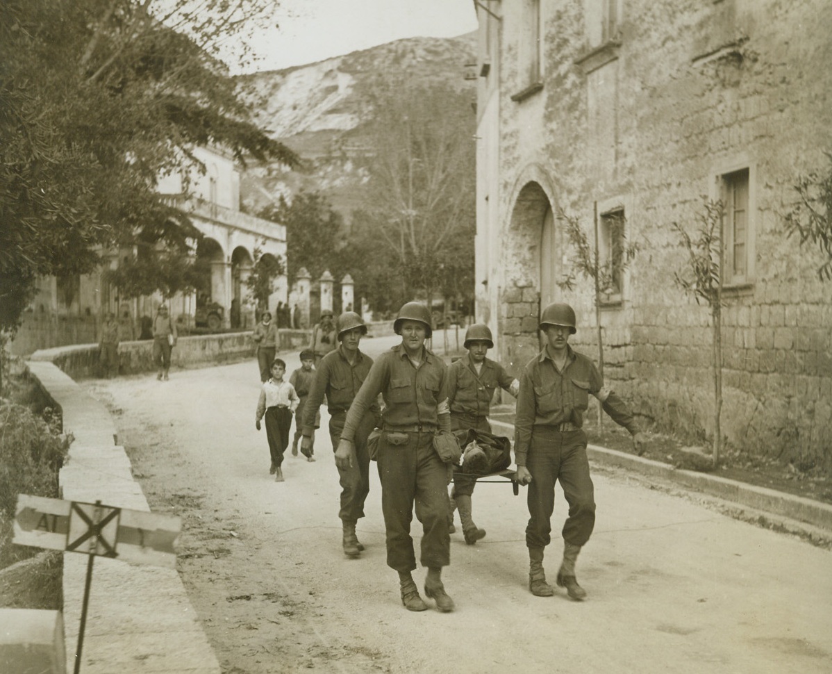 VOLTURNO CASUALTY, 10/24/1944. ITALY—American stretcher-bearers rush a casualty through the streets of a tiny Italian village, during the fight to cross the Volturno, and are followed by a pair of the town’s younger citizens. First aid has already been given to the wounded Allied fighter, and he is on his way to an ambulance.Credit: Acme photo by Bert Brandt, War Pool Photographer;