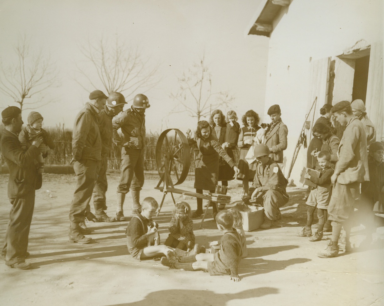 PEACE—IN THE HEART OF THE STORM, 2/13/1944. ON THE ITALIAN FRONT—Describing this photo Acme photographer Bert Brandt says: “This is the kind of scene you see as you travel along near the front—and it always amazes you for it looks so peaceful and yet you are within easy hearing of the shell bursts on the front only a mile away.” Left to right: Pfc Arthur Eames, Suffern, NY; Cpl Willie Whiteside, Rutherfordton, N.C.; and Cpl Arlo Pittman, May, Okla., watch with interest as an Italian family makes yarn from fleece in their farmyard just as they have done for years. Pvt Antone Silva, North Dartmouth, Mass., helps with the operation. Credit: Acme photo by Bert Brandt for the War Picture Pool;