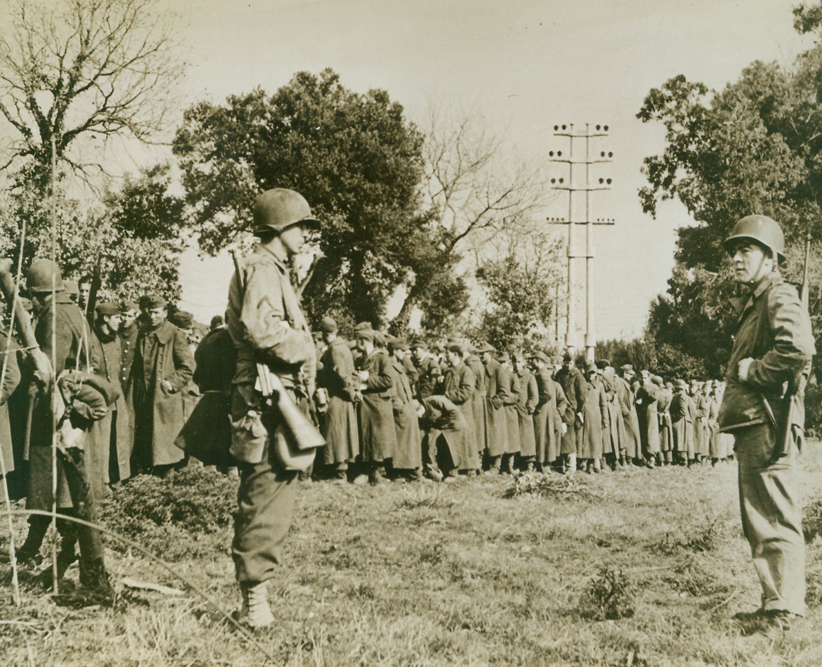 Nazis are the Fish, 2/11/1944. ITALY – German prisoners are herded together in the beachhead area south of Rome at a spot so wide-open the Yanks have nicknamed it the “Gold Fish Bowl.” Pvt. William Shea, (left), and Pfc. Andrew Calet, both of New York City, guard the Nazis who will be marched to the rear. Credit Line (Acme);