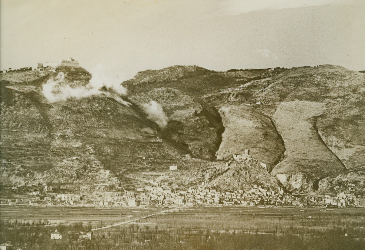 SMASHING NAZI DEFENSES AT CASSINO, 2/25/1944. ITALY—Allied shells burst between the town of Cassino and the famous Monte Cassino Abbey (left) on the tip of the mountain peak, as Fifth Army forces seek to drive the Germans from their strong defense positions. Other shells, which give off very little smoke and are barely discernible, are exploding in the town itself close to the castle which surmounts surrounding houses. Credit: Acme;