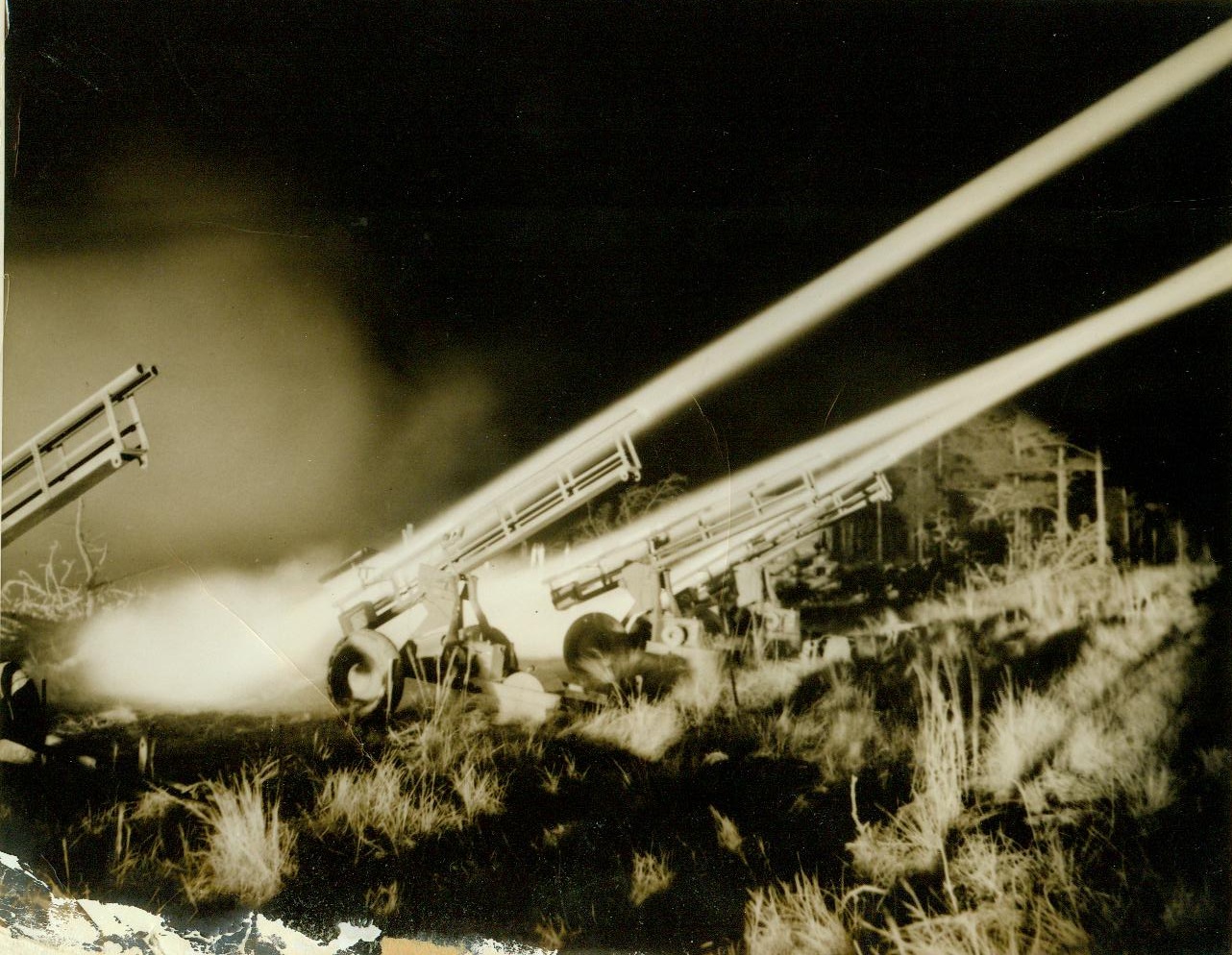 ROCKET TARGETS FOR ACK-ACK TRAINING, 3/8/1944. CAMP DAVIS, N.C. – Jet-propelled rocket targets are now being used to train anti-aircraft artillerymen at Camp Davis, giving our gunners practical experience in firing at “enemy planes” diving at terrific speed, and preparing them for action against the jet-propelled planes of tomorrow, should the enemy perfect such planes before the war is over. Fired from specially designed carriages, the rockets have an initial velocity of 450 miles per hour or 675 feet per second. They are released from the range at Holly Shelter, a Camp Davis firing point. Manning 50 caliber machine guns and 20 and 40mm weapons, the trainees improve their aim and accuracy as they track the fast-moving, 59-inch targets. The rocket always describes an approximate parabola and has a maximum horizontal range of about 2200 yards. The following photos show phases of rocket target practice by night and by day. NEW YORK BUREAU Here is a closeup of the launching carriages as the rockets zoom out of them. Leaving a cloud of bright dust on the ground behind the carriages, the targets cut a broad band of light in the sky. Credit: ACME;