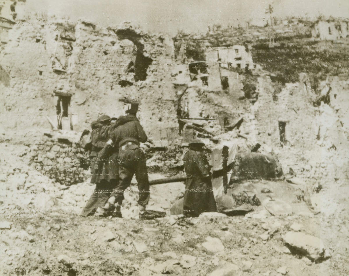 Wounded Return, 3/31/1944. CASSINO, ITALY—With the flag of the Red Cross waving protectively before them, wounded New Zealand warriors are helped back from the fighting lines in Cassino area. With one arm slung over the shoulder of each medical corpsman helping him, one of the fallen fighters stumbles through the ruined sector.  Credit:  OWI radiophoto from ACME.;