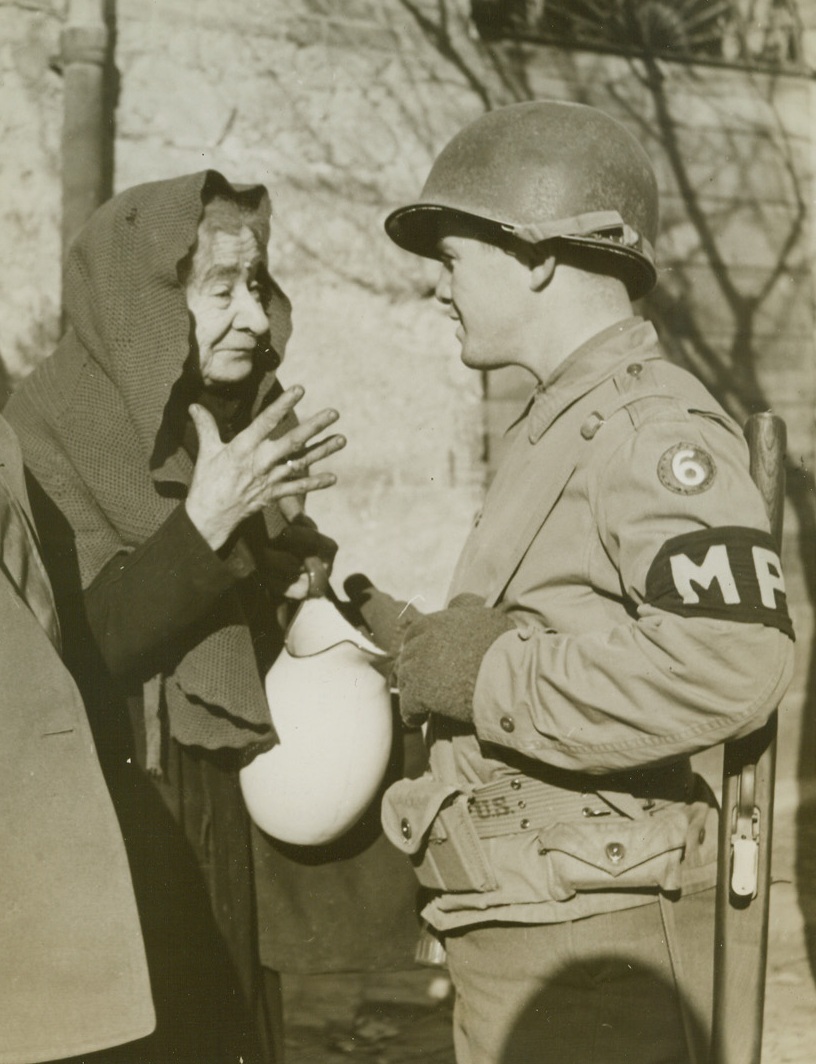 “Most Kissed Woman”,  3/11/1944. NETTUNO-ANZIO BEACHHEAD – Unlike other Italian towns, where beautiful signorinas waited to greet advancing Yanks with kisses, the Nettuno-Anzio beachhead was barren of feminine attraction, except for 81-year-old Signora Marcella Giorgi. One of the few women permitted to remain behind (because she must care for her invalid husband) when other civilians were evacuated from the area, she’s a one-woman welcoming committee from Allied fighting men. She insists on kissing every arriving soldier by way of greeting. Signora Giorgi is shown here chatting with Pfc. George W. DeNicola. Credit Line (Acme photo by Charles Seawood for the War Picture Pool) WP;