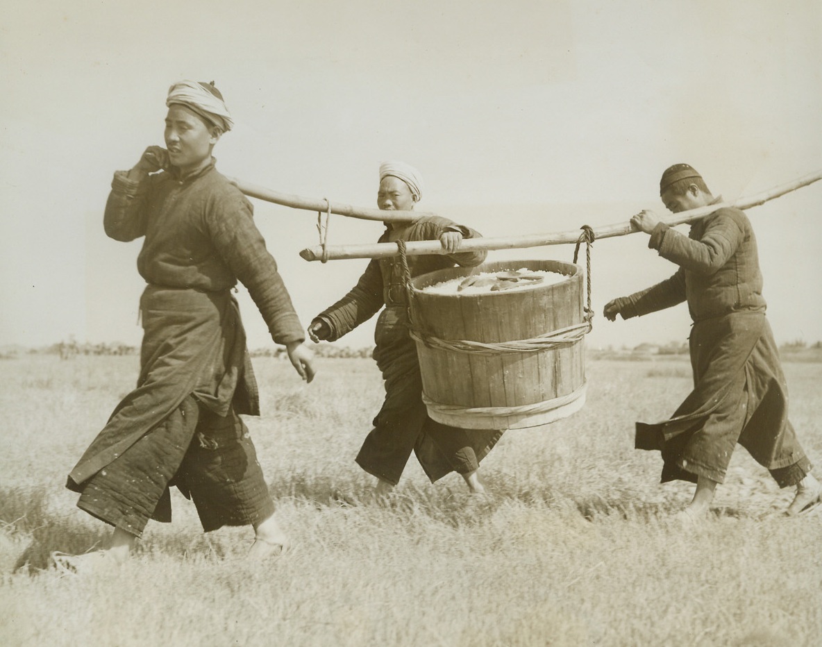 Airfield-Handmade, 3/31/1944. China – Lack of modern equipment and machinery isn’t stopping resourceful Chinese from building modern airfields in China. Three hundred thousand Chinese workers have been conscripted to do the job. A hundred thousand little handmade wheelbarrows are used to transport materials – and even the chipping of stones for the runways is done by hand. American Engineers are supervising the work, in conjunction with expert Chinese Army Engineers Photo shows: The laborers eat their meals out on the runway so that no time is lost going to and from lunch. This Chinese K.P. outfit carries a bucket of rice out to the workers. Credit: ACME photo by Frank Cancellare for the War Picture Pool;