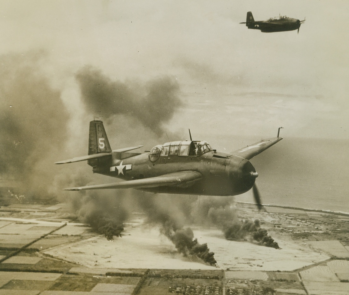 Jap Planes Blasted in Marianas, 3/10/1944. Tinian Island  Huge pillars of smoke rise from direct hits on Jap planes on Tinian island airfield as a result of attack by TBF Avengers in the Marianas.  Our planes breeze away from their destructive work.  Square patterns in foreground are sugar cane fields adjoining the enemy airstrip. Credit line (ACME);