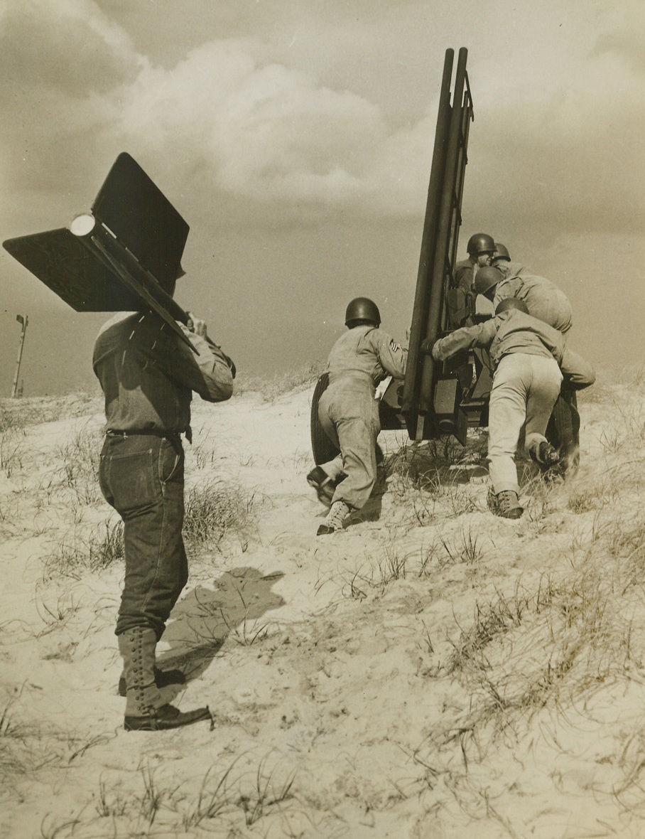 Rocket Targets for Ack-Ack Traininga, 3/8/1944. Camp Davis, N.C.—Jet-propelled rocket targets are now being used to train anti-aircraft artillerymen at Camp Davis, giving our gunners practical experience at firing at “enemy planes” diving at terrific speed, and preparing them for action against the jet-propelled planes of tomorrow, should the enemy perfect such planes before the war is over. Fired from specially designed carriages, the rockets have an initial velocity of 450 miles per hour or 675 feet per second. They are released from the range at Holly Shelter, a Camp Davis firing point. Manning 50 caliber machine guns and 20 and 40 mm weapons, the trainees improve their aim and accuracy as they track the fast-moving, 59-inch targets. The rocket always describes an approximate parabola and has a maximum horizontal range of about 2200 yards. The following photos show phases of rocket target practice by night and by day.New York BureauWhile other members of the rocket-launching crew push the specially designed carriage into place at Holly Shelter, another crewman carries the heavy, triple-finned rocket on his shoulder. The rockets are painted white, when fired at night, to increase visibility.Credit: ACME.;