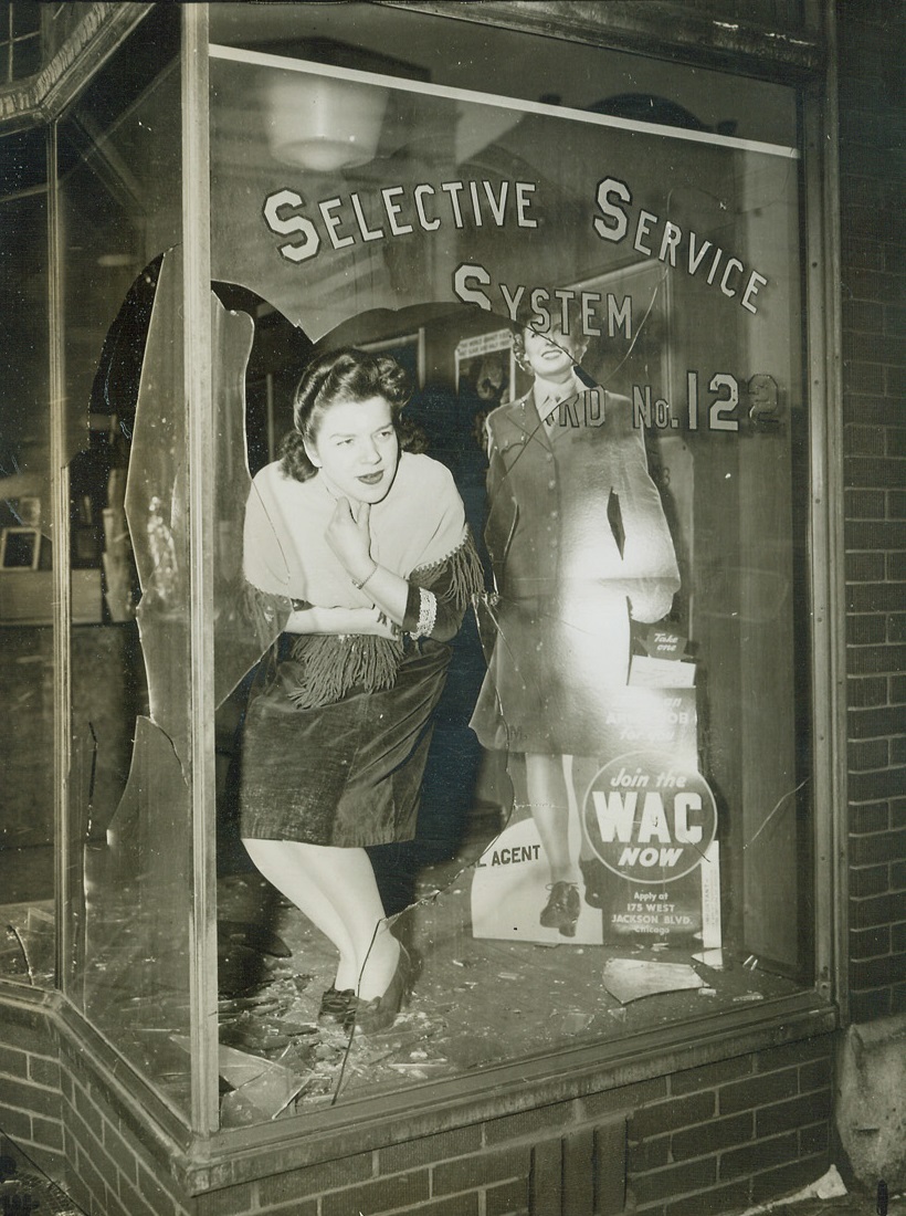 Draft Office Smashed, 3/8/1944.Chicago – Six huge plate glass windows and a glass door of the office of Selective Service Board No. 122 in Chicago were smashed by an early morning marauder whom neighbors reported drove a plant through the glass. Officials blamed this, the second attack, on “discontented people who don’t want to go into service or see their friends go.” Here, Theresa Munari, a draft board clerk, inspects the damage. Credit: ACME;