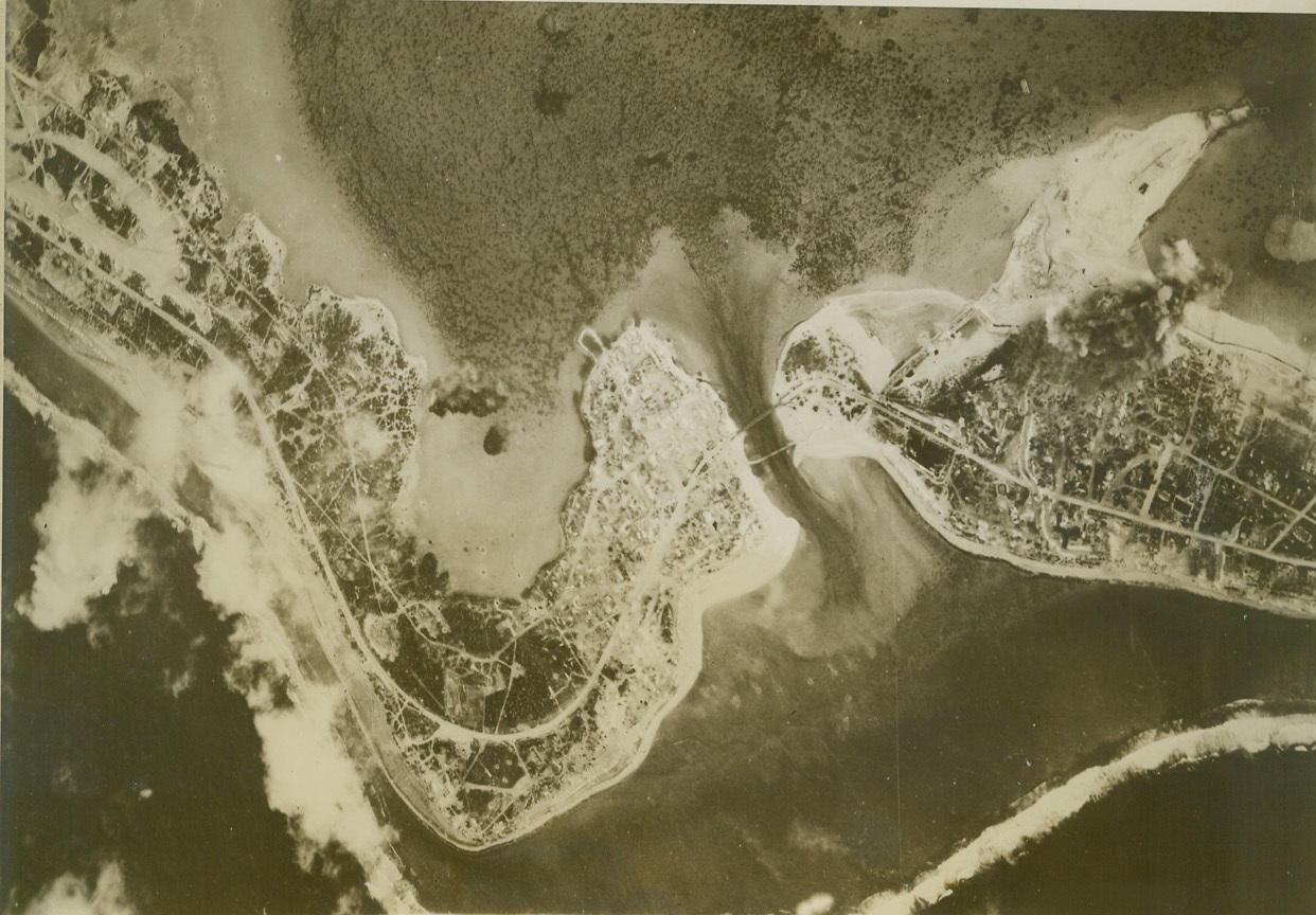 WAKE ISLAND BLASTED AGAIN, 5/2/1944. WAKE ISLAND – Huge clouds of smoke rise from Wake Island when 7th AAF bombs hit the area in which most of Jap construction is concentrated. This attack, made about a week ago, shows Jap efforts to strengthen the island. Numerous place revetments – all empty- are shown at the end of the main runway. Necklace of white dots on the beach are piles of sand made by Japs as anti-tank traps against constant threat of U.S. invasion. Credit: ACME;