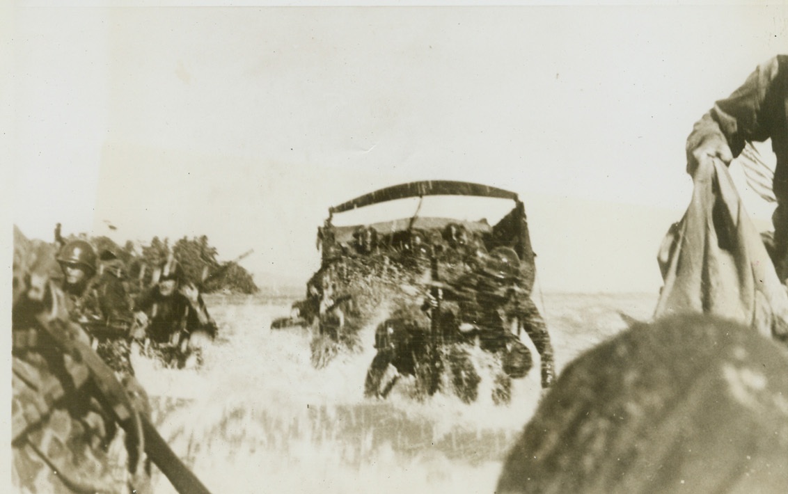 FIRST AID FOR AN INVADER – 1, 6/1/1944. WAKDE ISLAND – As the second wave of infantrymen charge ashore in the assault on Wakde Island, one of the invaders slips to his knees in the surf, hit by Jap machine gun fire. This unusual series showing on-the-spot first aid for a fighting man was made by 1st Lt. Sidney Simon, who was in on the Wakde Island invasion as an observer for the U.S. Army Engineers. Under enemy fire every moment, Lt. Simon’s camera recorded the heroic mercy treatment rendered a fallen comrade in the midst of landing operations. Credit: Photo by Lt. Sidney Simon from ACME;