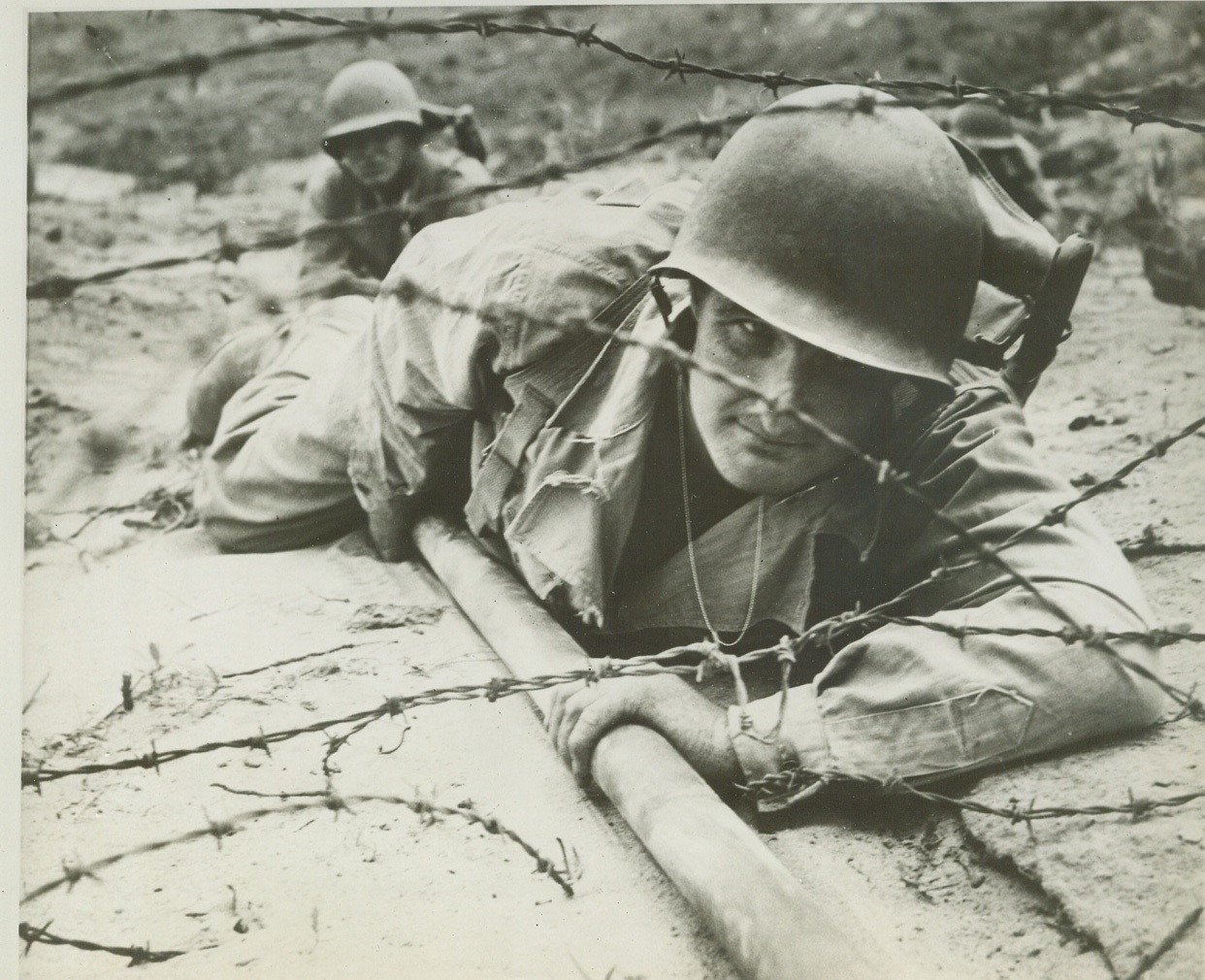 The Infantry School's "West Wall", 6/4/1944. Ft. Benning, Ga. – Newest assault course at Fort Benning is the infantry school’s authentic exterior replica of a section of Germany’s famed West Wall, a series of pillboxes and blockhouses, which extends along the coastline from France to Norway and which our invasion troops must reduce from beachheads before they can drive inland to strike at the heart of the Nazi-dom. Constructed after study from Allied photographs, the course covers 1,000 square yards and is used by the infantry school in the training of officers and officer candidates. Infantry school demonstration troops, some of whom are shown in these photos, carry out “assaults” for the benefit of each new class. Live ammunition is used and every three weeks the concrete pillboxes and blockhouses must be largely rebuilt and barbed wire torn by Bangalore torpedoes must be restrung by engineer troops assigned to the school. PHOTO NO. 1: First step in the reduction of a pillbox or blockhouse, as taught by the infantry school, is the blasting of a path though barbed wire. This is accomplished by use of the British-invented Bangalore torpedo, which opens the way for the flame-thrower. Here demonstration troops place the long torpedo under the wire. Credit Exclusive “ACME” Photo;