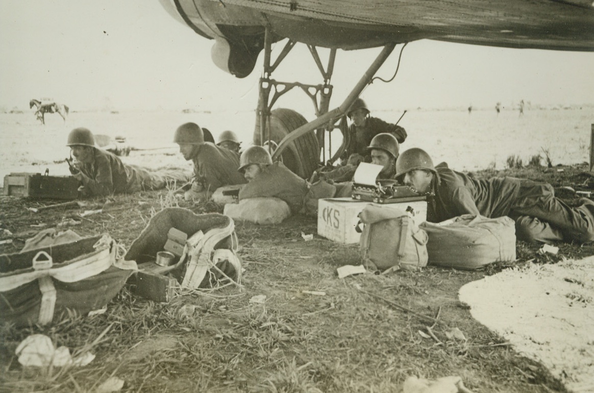 Wing of Plane Offers Refuge from Strafing, 6/17/1944. Makyina, Burma – American Soldiers at Makyina Airdrome, which was taken from the Japs by Merrill’s Marauders, take refuge under the wing of a C-47 as a Jap sniper bullet whizzes by. After the capture of the field, the American and Chinese soldiers were constantly bothered by Jap snipers and strafing planes. Note the correspondent with paper in his typewriter, ready to bang out a story, or else caught in the middle of one. Credit: ACME;