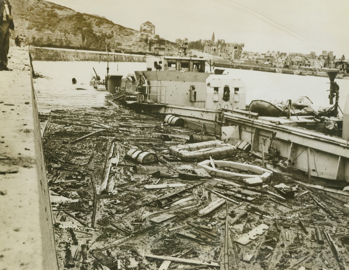 Remains of German Flak Boat, 6/15/1944. France – Amid the floating debris of war, a German flak boat lies where troops of the Armies of Liberation found her, in the harbor at Port-en-Ressin, France. Blasted buildings along the shore show evidence of the power of the naval barrages that supported the invasion troops. Credit: U.S. Navy photo from Acme;