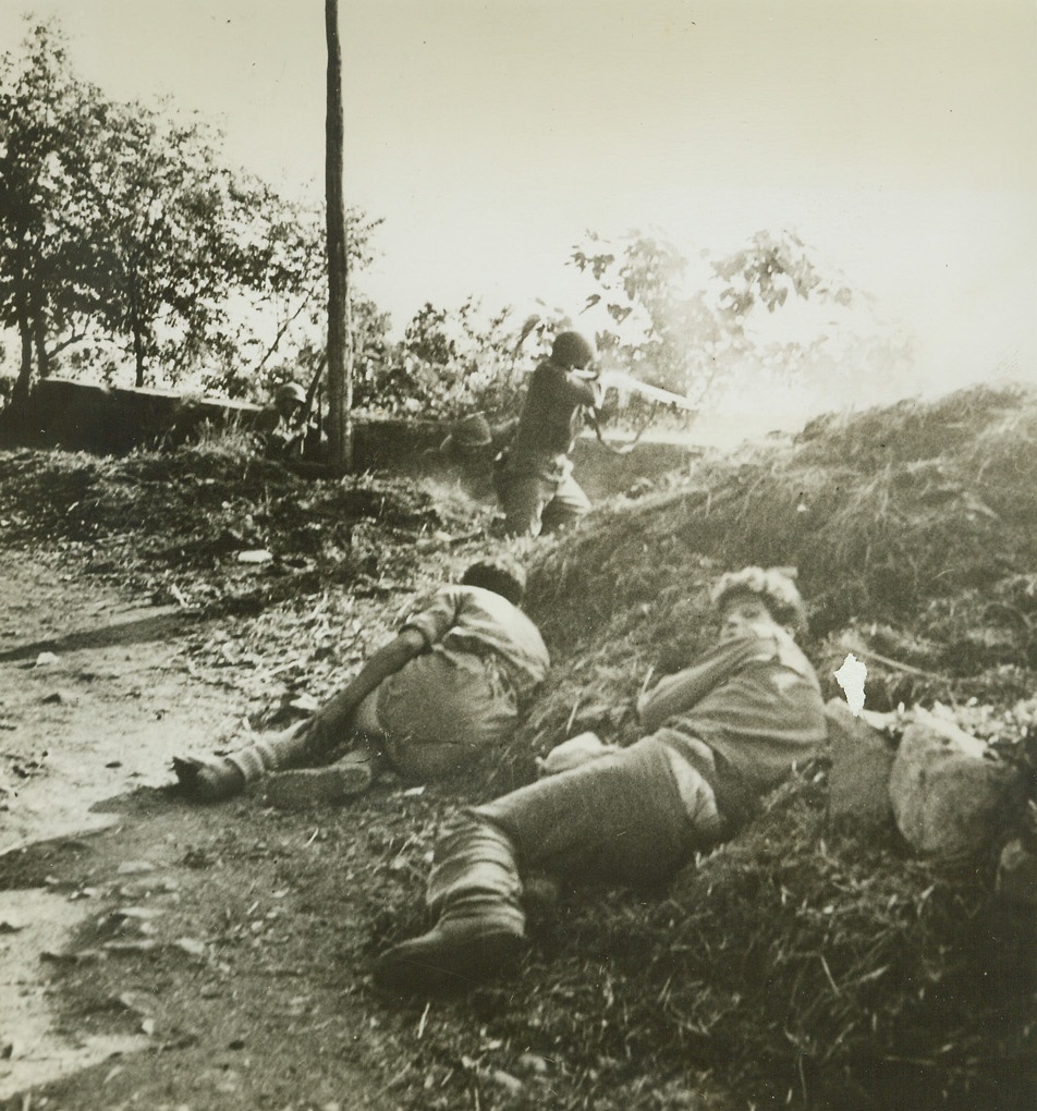 Yanks in a Spot – 5, 7/14/1944. Nemi Valley, Italy – Once the peaceful site of fragrant vineyards and the center of a flourishing wine-grape business, beautiful Nemi Valley, near Rome, was turned into a hot spot recently when a scouting patrol of Yank Infantrymen, guided by Italian partisans, found German strength much greater than they had suspected. Our boys had to fight their way out through holes in a wall of enemy machine gun fire. Here, Italians who led the fighters to concealed German positions in the valley crouch low behind a pile of debris as the Yanks send bullets screaming into enemy positions. Credit: Yank Magazine photo from ACME;