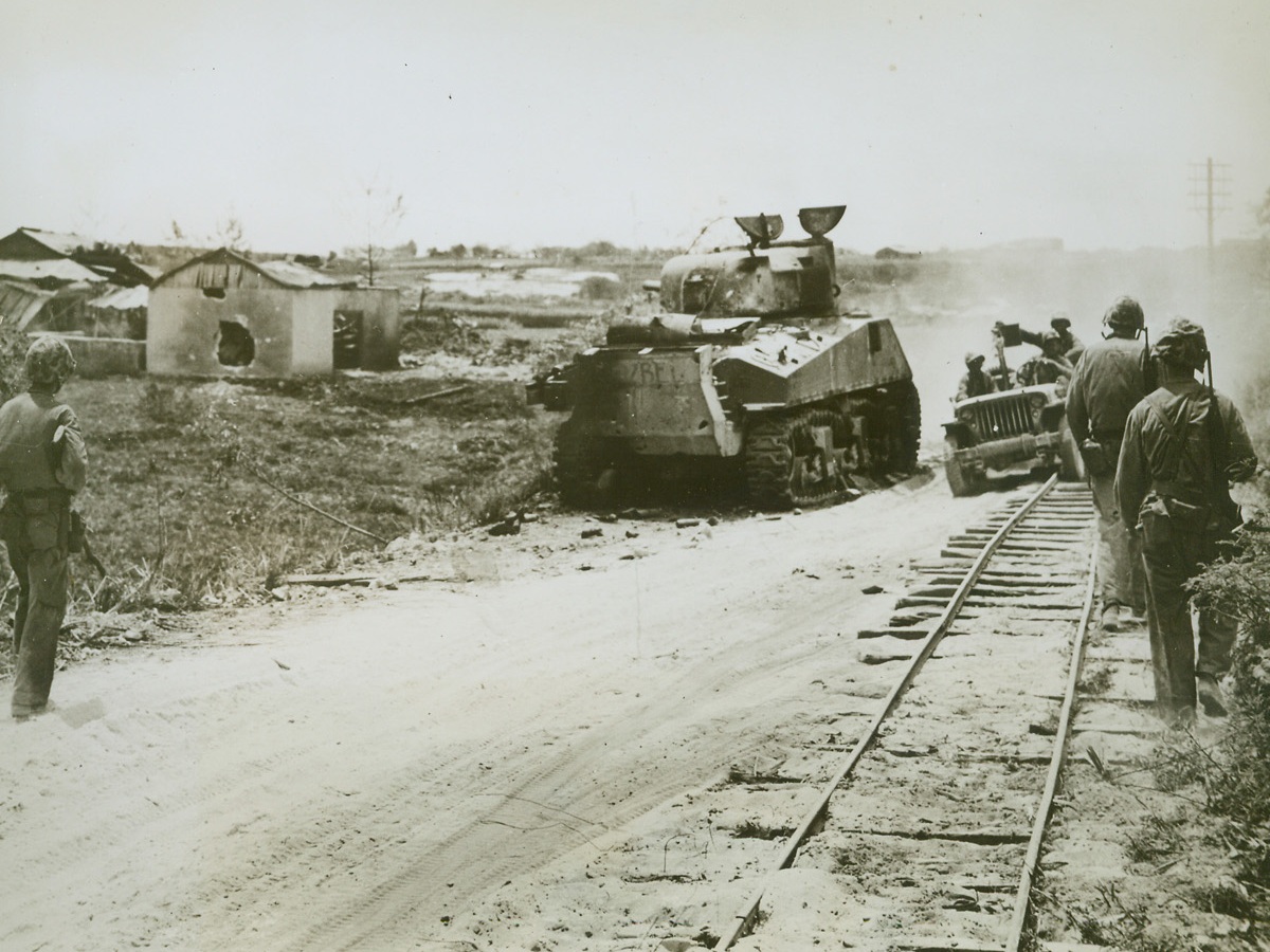 Stilled Scene of Battle, 7/1/1944. Saipan – Marines on foot and in a jeep, bypass the charred remains of an American tank along a road on Saipan.  The Leatherneck tankmen fought a battle with Japs hidden in the concrete building at which the turret gun is pointing.  A gaping hole in the side wall is evidence of a direct hit by one of the shells from the tank. Credit (Marine Corps photo from ACME);