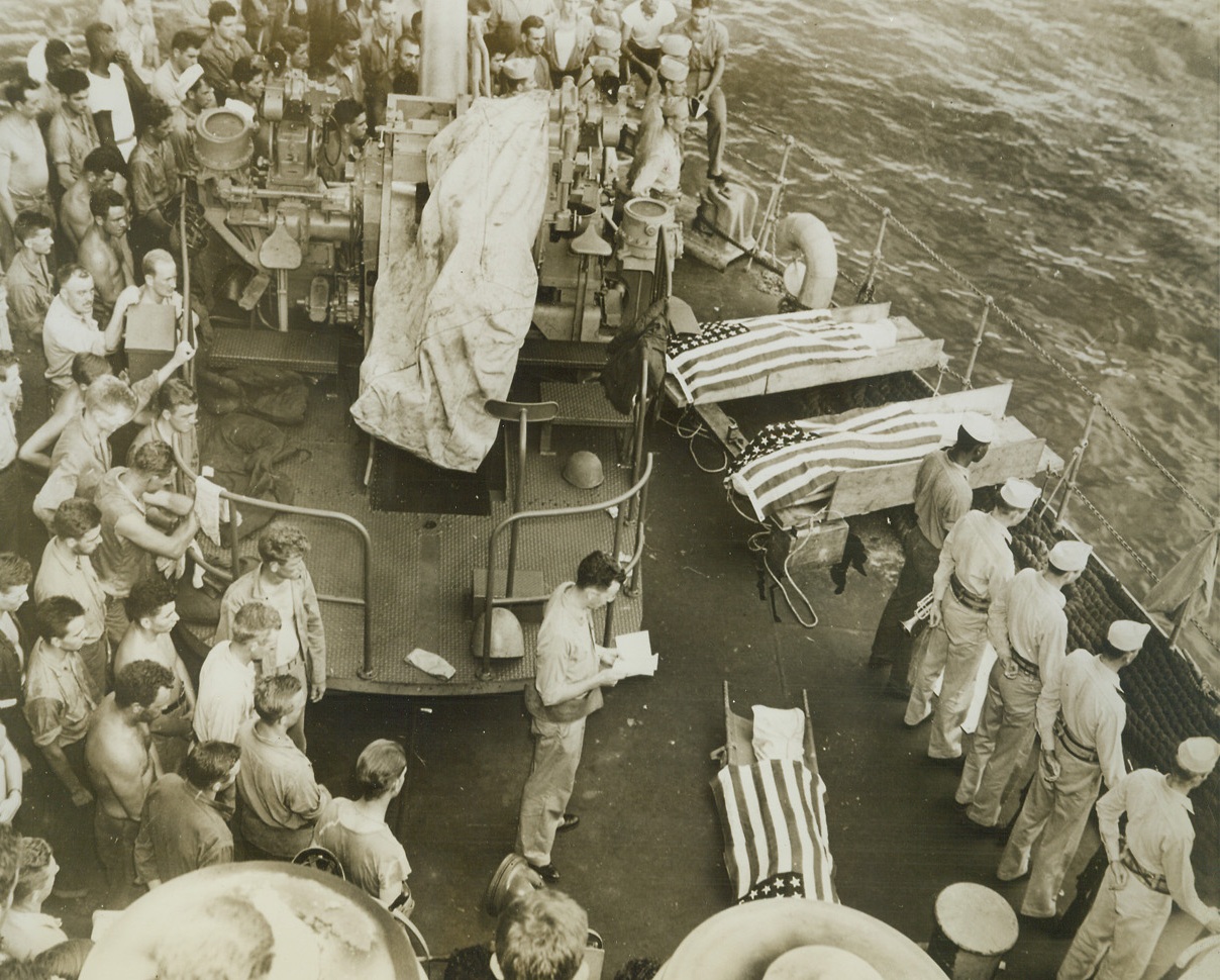 SEA GRAVE FOR AMERICAN HEROES, 7/7/1944. SAIPAN—While the noise of battle rumbles in the distant, three Americans who gave their lives in the early assault waves on Saipan are buried at sea from a Coast-Guard-manned transport. As the chaplain reads the simple service, Coast Guardsmen stand solemnly near the flag-draped bodies. When the honored dead were consigned to the sea, the guard of honor (lower right) fired a volley and taps was sounded. Scene took place off Saipan on the third day of the invasion.  Credit: COAST GUARD PHOTO FROM ACME;