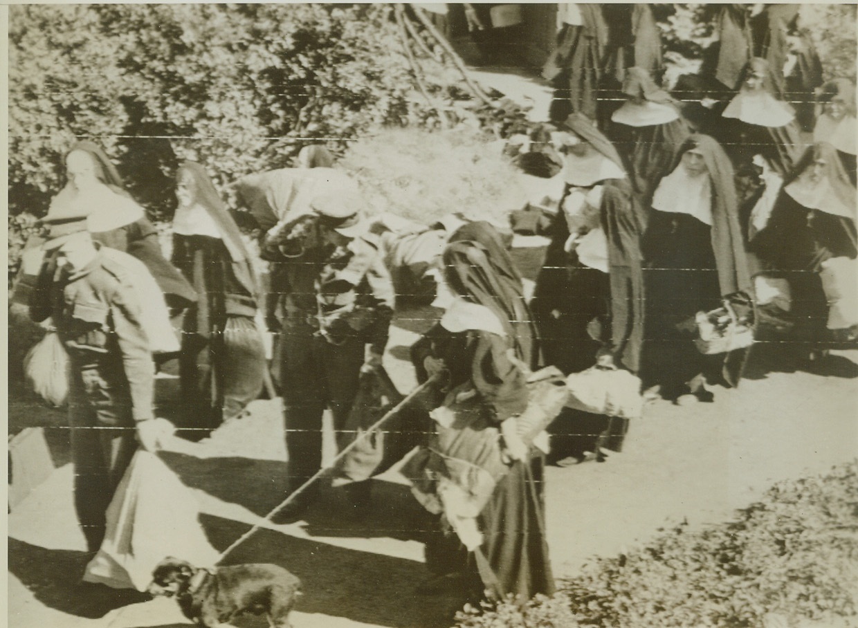 Transported to Safety, 7/16/1944. Bayeux, France—RAF officers carry the luggage of nuns who were transported to liberated Bayeux from the fighting fronts in Normandy. Nearly 100 Benedictine sisters were evacuated from Caen and surrounding areas where fighting was in progress. One of the sisters leads a little dachshund.  Credit: British official photo via U.S. Signal Corps radiotelephoto from ACME;