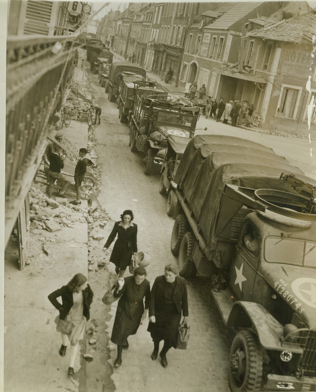 Common Sight, 7/16/1944. Isigny, France—No longer fascinated by the endless column of war supplies woving through their town, for the sight has become a common one by now, natives of Isigny go about their business ignoring the convoy passing beside them. The heavy trucks, on their way to the embattled St. Lo sector, drive slowly through the narrow streets lined with buildings that still bear the scars of war.  Credit: ACME photo by Bert Brandt, for the War Picture Pool;