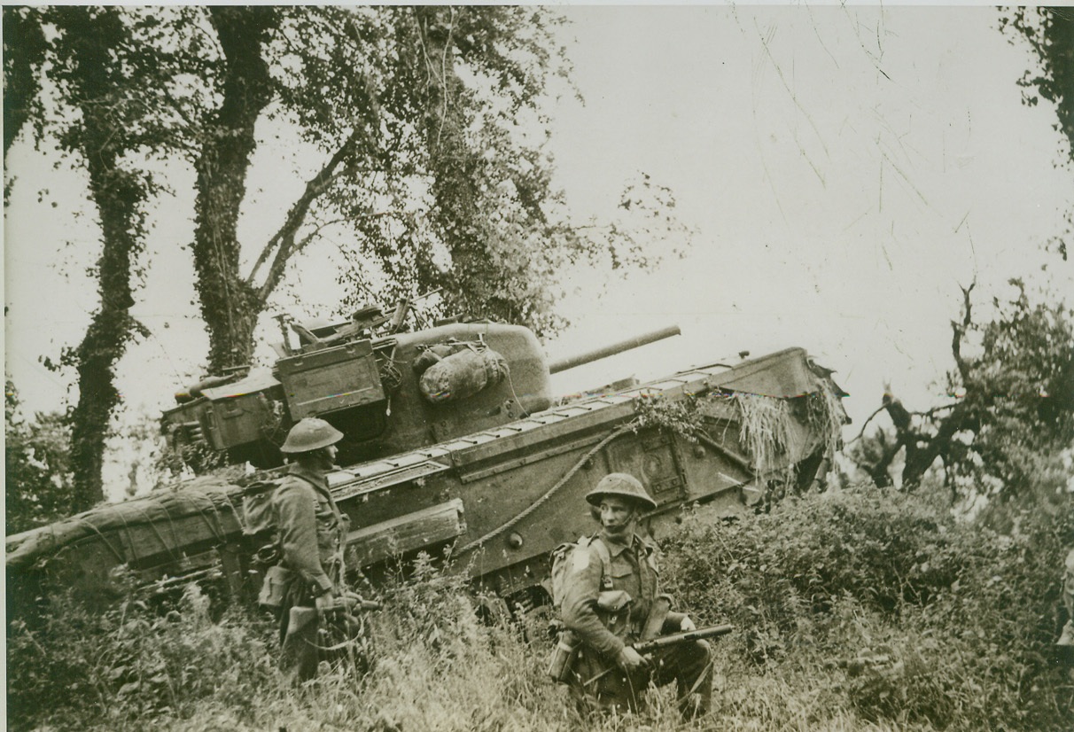 British Move Forward In Caen Sector, 7/4/1944. Caen, France—A lumbering Churchill tank crashes through a hedge at the side of a field, where British troops wait to follow under cover of the armored vehicle. Today (July 4) British and Canadian forces captured Carpiquet, only 3 miles west of Caen, and endangered German troops holding a semi-circular line north of the town. Credit: British official photo from ACME;