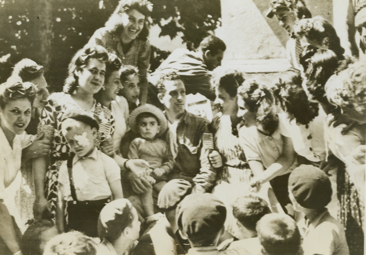 A RIGHT WELCOME WELCOME, 8/22/1944. FRANCE – Completely surrounded by lovely girls and curious French tots, an American soldier seems embarrassed?  At the situation he is in.  Scene took place far inland from the South coast of France at Riez, where the entire population greeting passing American troops driving Northward beyond the city.Credit: Army radio telephoto from Acme;