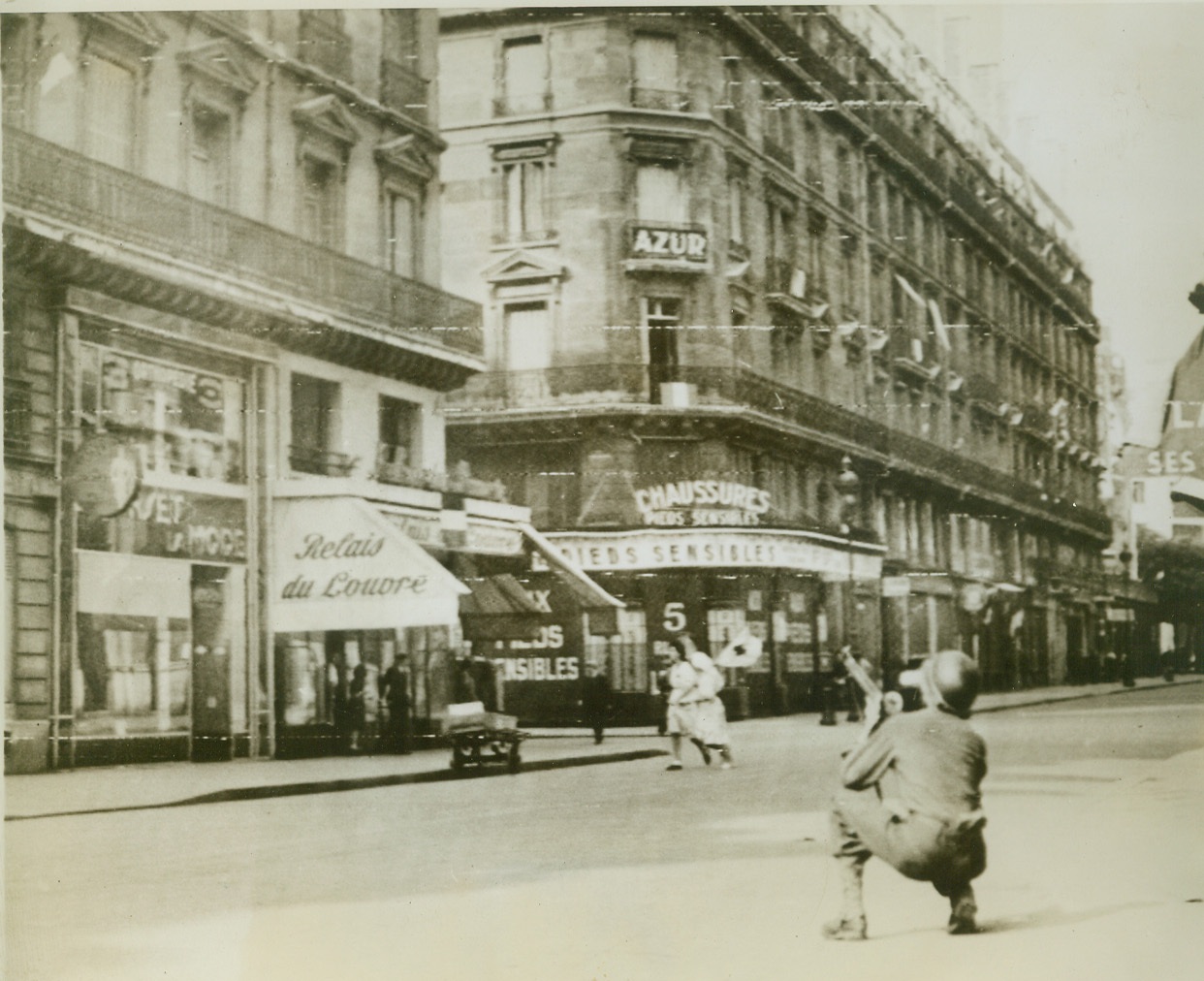 Snipe-Hunting in Paris Streets, 8/28/1944. Paris—As two French Red Cross nurses scurry for safety from a sniper who opened fire from a building in Paris, an American infantryman, with no shelter, kneels in the street and seeks to locate the sniper. Credit: Signal Corps radiotelephoto from ACME;