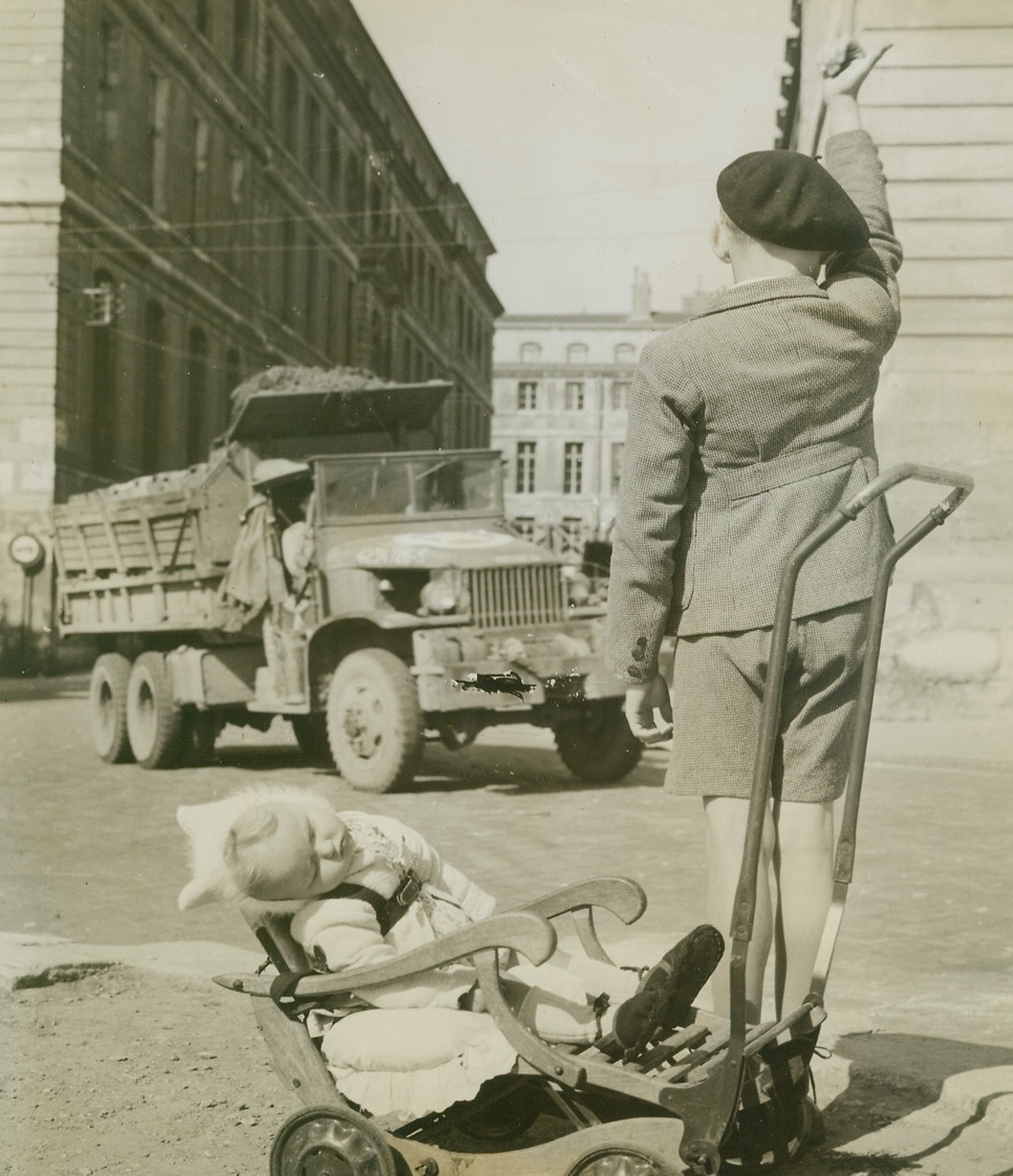 GOING BYE-BYE?, 9/24/1944. PARIS—Catching on quick to the customs of “Les Yanks”, this young Parisian hoists an optimistic thumb into the air as an Army truck lumbers by. Knowing that American soldiers are “softies” with kids, he’s hoping for a lift back home for himself and his sleeping baby sister. Photo by Charles Haacker, Acme photographer for the War Picture Pool.Credit: Acme;