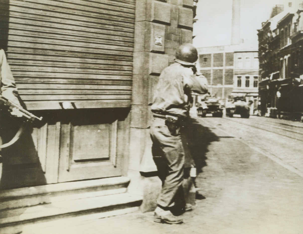 German Sniper His Target, 9/13/1944. BELGIUM—A Yank infantryman pokes the muzzle of his rifle around a street corner in Liege, Belgium, to take a shot at a German sniper hidden in a building at the end of the street. Allied armored vehicles rumble up the road. Credit: ARMY RADIOTELEPHOTO FROM ACME.;