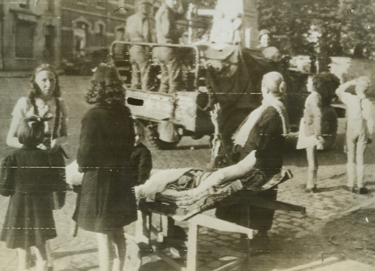 Belgian Children Recognize Yanks as Liberators, 9/14/1944. BASTOGNE, BELGIUM—Wheeled out onto the street on her rolling cot, a little invalided Belgian girl waves to American soldiers as the Yanks enter Bastogne in the wake of retreating Germans. To the right, a little boy snaps to attention and gives a smart salute—American style. Credit: SIGNAL CORPS RADIOTELEPHOTO FROM ACME;