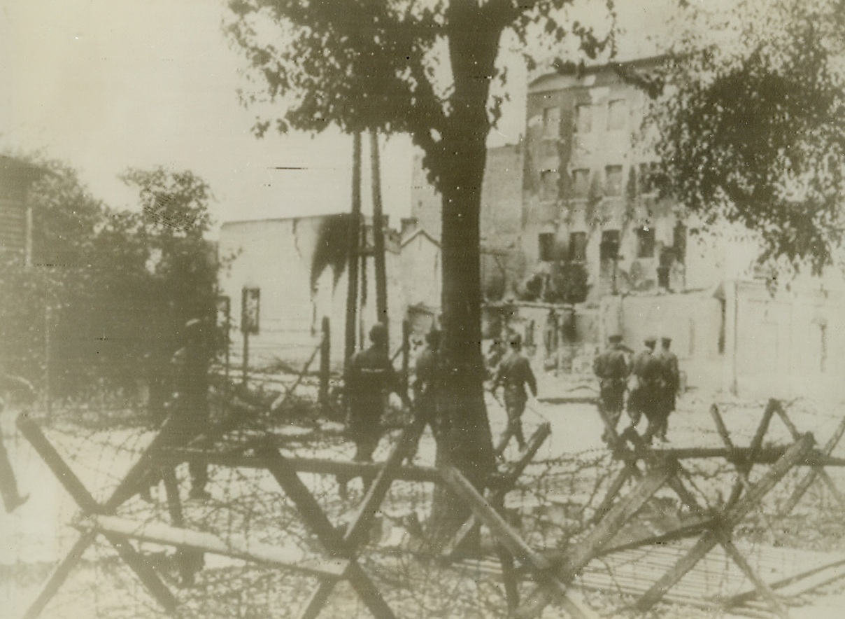 Polish Patriots Revolt in Warsaw, 9/2/1944. According to the German caption on this photo, radioed today (September 2) from Stockholm, German soldiers march past barbed wire barricades as they seek Polish patriots who are participating in the revolt within Poland’s capital. As the fighting continues, the Polish government appeals to the Allies for aid in wrestling the capital from the Nazis.Credit (Acme Radiophoto);