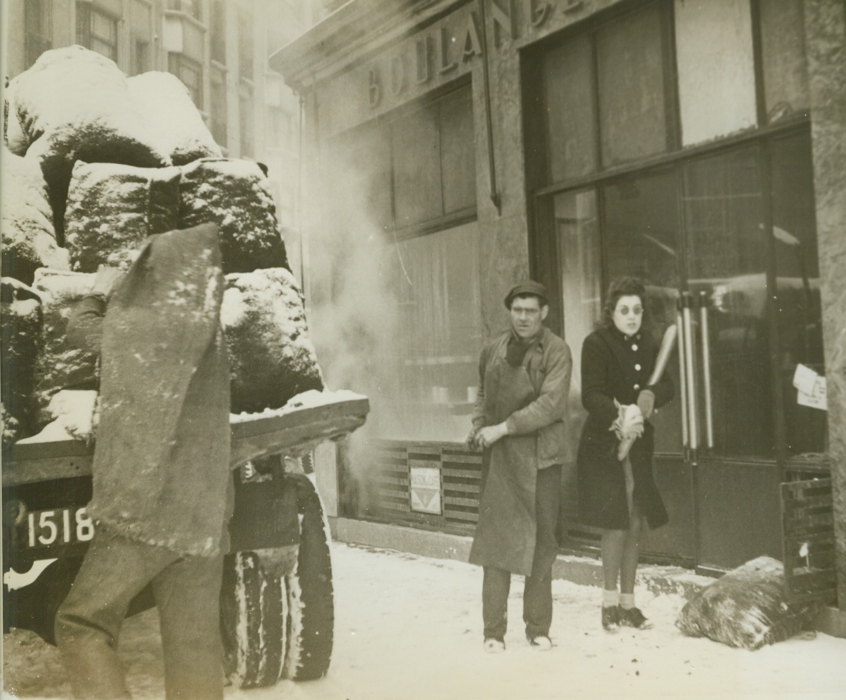 In Paris, It’s Cold and Food Is Scarce, 1/20/1945. Paris -- Parisians have had a tough time keeping warm this winter and almost as tough a time getting their daily ration of food. Homes, offices, style salons, all have a near constant temperature of near zero centigrade and no relief is in sight. Warm clothing is scarce, coal is nonexistent even in the black market and the small quantities of firewood on hand are hard to get and prohibitive in price. As for food, French officials say the situation is “normal” after four years of occupation, but all food supplies are very limited. This series of photos, by Alan Montrose for the War Picture Pool, shows what Parisians are enduring this very cold winter. New York Bureau There is some coal in Paris but it goes to hospitals and to baker’s ovens to provide necessary rations of bread for the none-too-well-fed people of the city. This coal is being unloaded in front of a bakery shop. A woman, shivering against the cold, clutches her all-important loaf. Credit: ACME;