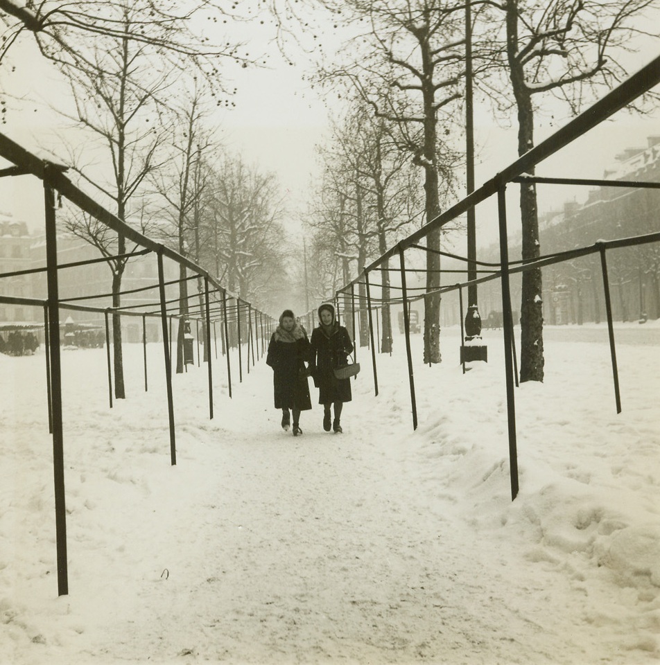 In Paris, It’s Cold and Food Is Scarce, 1/20/1945. Paris -- Parisians have had a tough time keeping warm this winter and almost as tough a time getting their daily ration of food. Homes, offices, style salons, all have a near constant temperature of near zero centigrade and no relief is in sight. Warm clothing is scarce, coal is nonexistent even in the black market and the small quantities of firewood on hand are hard to get and prohibitive in price. As for food, French officials say the situation is “normal” after four years of occupation, but all food supplies are very limited. This series of photos, by Alan Montrose for the War Picture Pool, shows what Parisians are enduring this very cold winter. New York Bureau Two heavily-clothed women walk past empty booths in one of Paris’ largest open-air shopping districts. This was an “open” day but because of food shortage and a possible heavy snowfall halting deliveries, a majority of the stalls were empty. In background at left crowds gather round the few stalls that had enough foodstuffs to open for business. Credit: ACME;